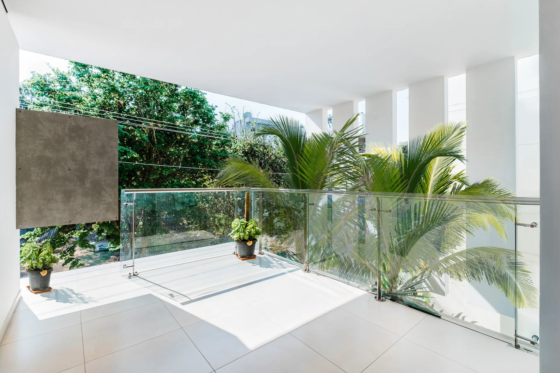 A modern white balcony features large tile flooring, a clear glass railing, potted plants, and a view of lush green trees.