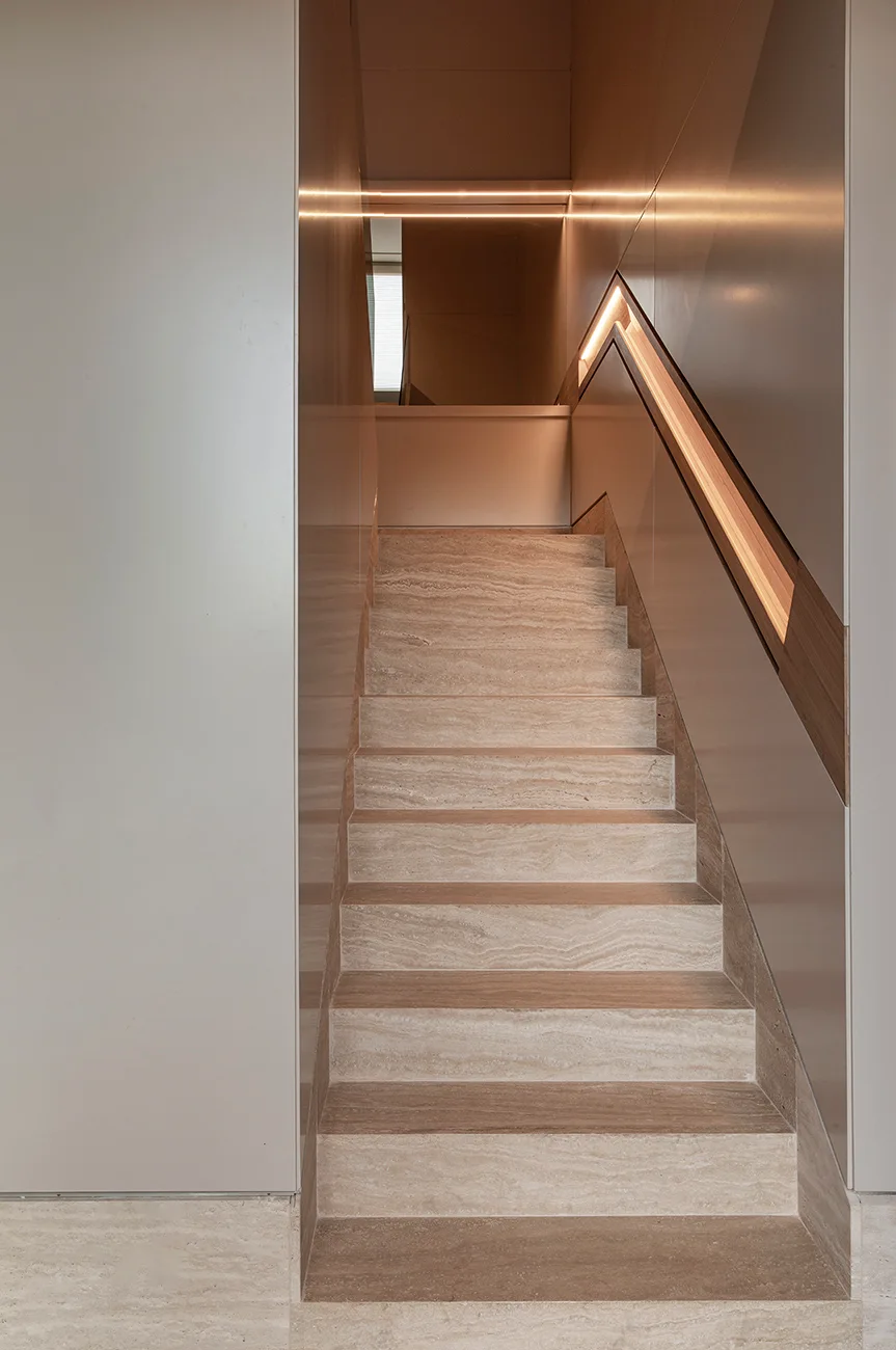 A minimalist beige stone staircase features smooth wall panels and a recessed wooden handrail illuminated by an integrated LED strip light.