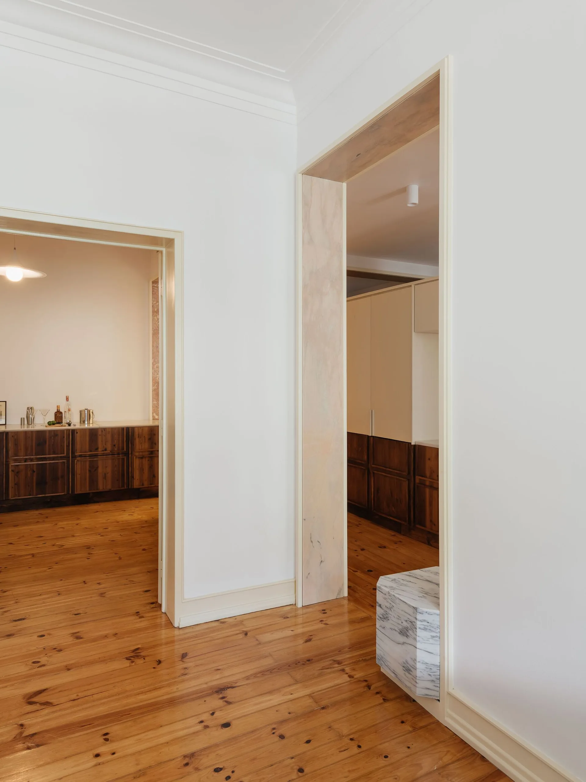 A bright hallway with pine wood floors features white walls and two doorways, one lined with veined marble next to a small marble plinth.