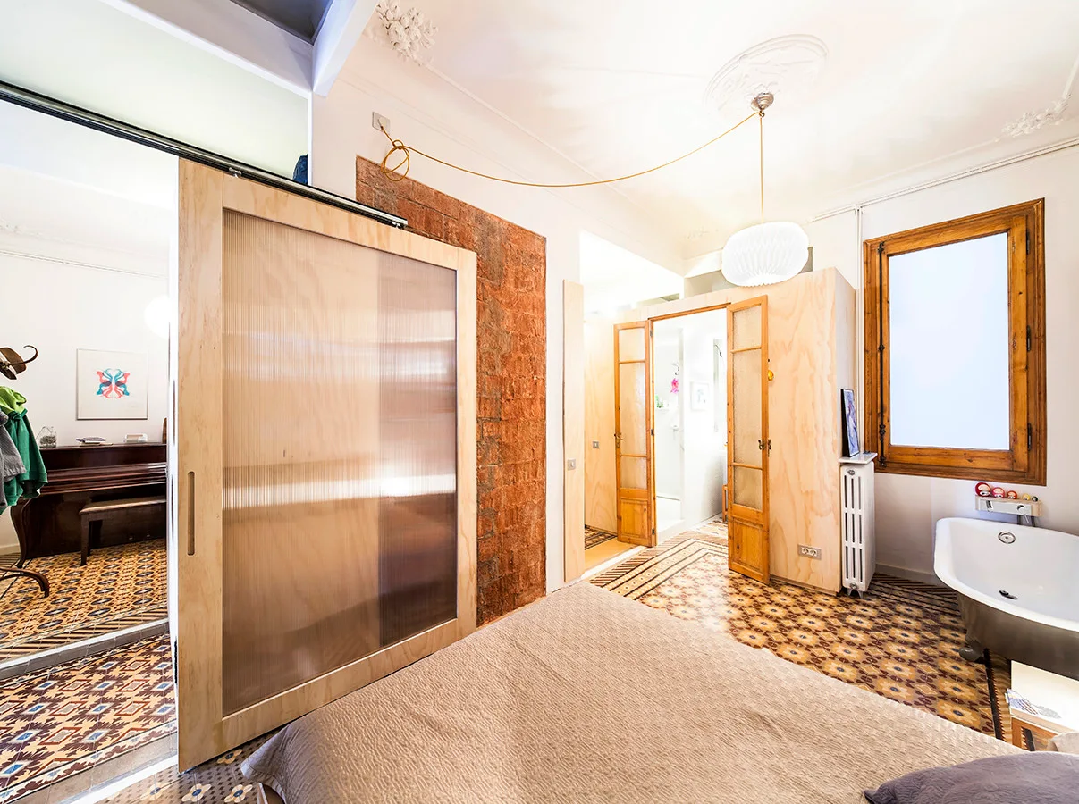 An eclectic bedroom featuring a custom plywood sliding door, patterned floor tiles, an exposed brick accent, and a white clawfoot bathtub.