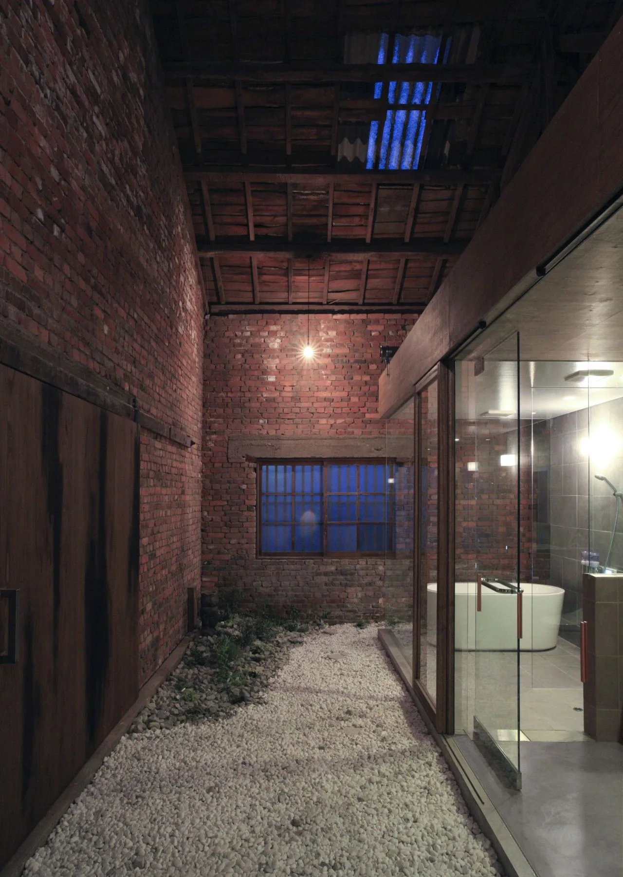 An indoor glass-enclosed bathroom with a white bathtub sits next to a white gravel path under exposed brick walls and a timber ceiling.