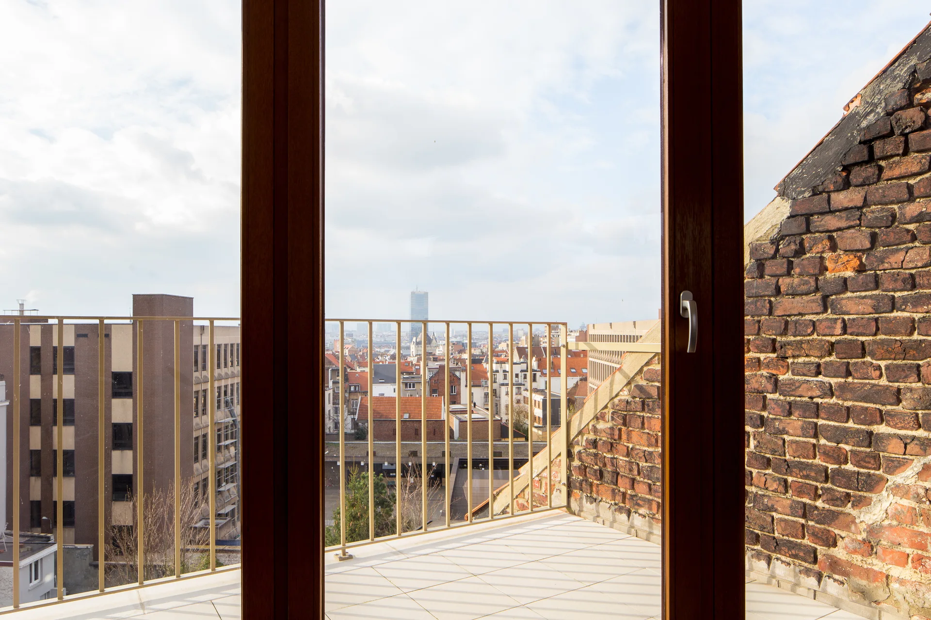 A view through a dark wood window frame onto a tiled balcony with a weathered brick wall and a metal railing overlooking a cityscape.