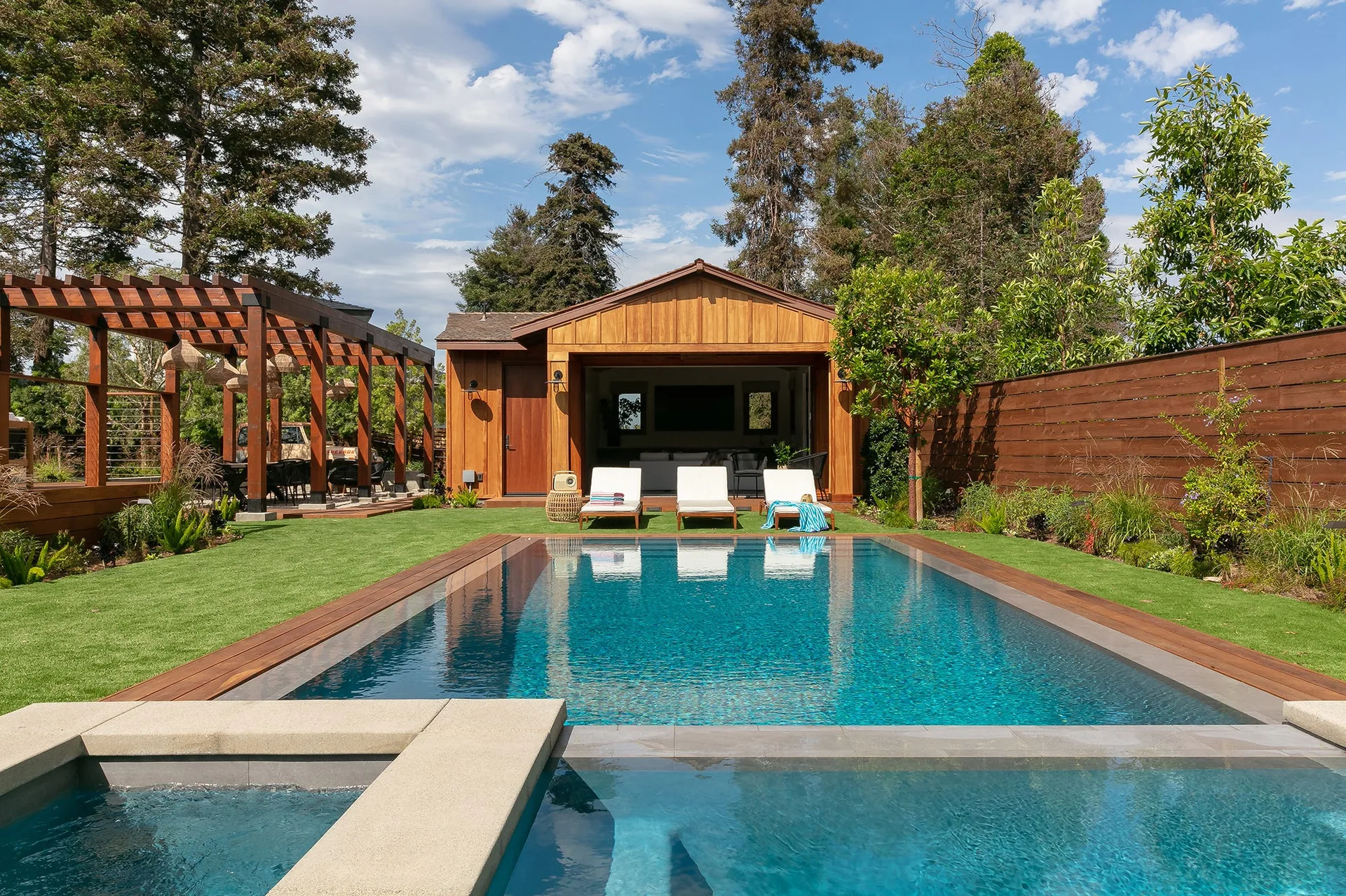A rectangular swimming pool is surrounded by a green grass lawn, white lounge chairs, and a warm wood pool house under a blue sky.