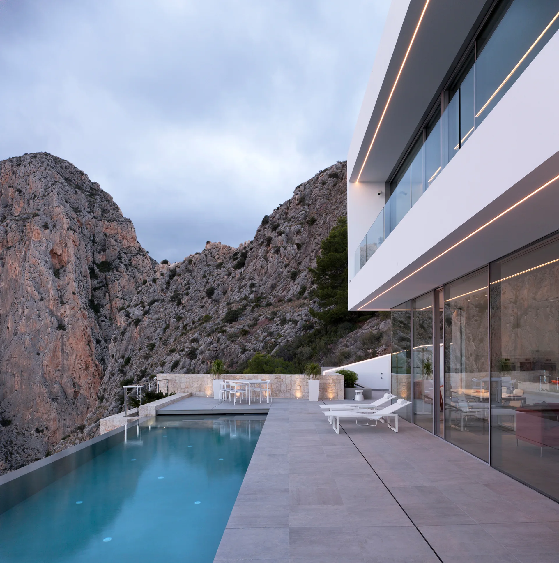 A modern white home with large glass walls overlooks an infinity pool and grey tiled terrace set against a steep mountain canyon.