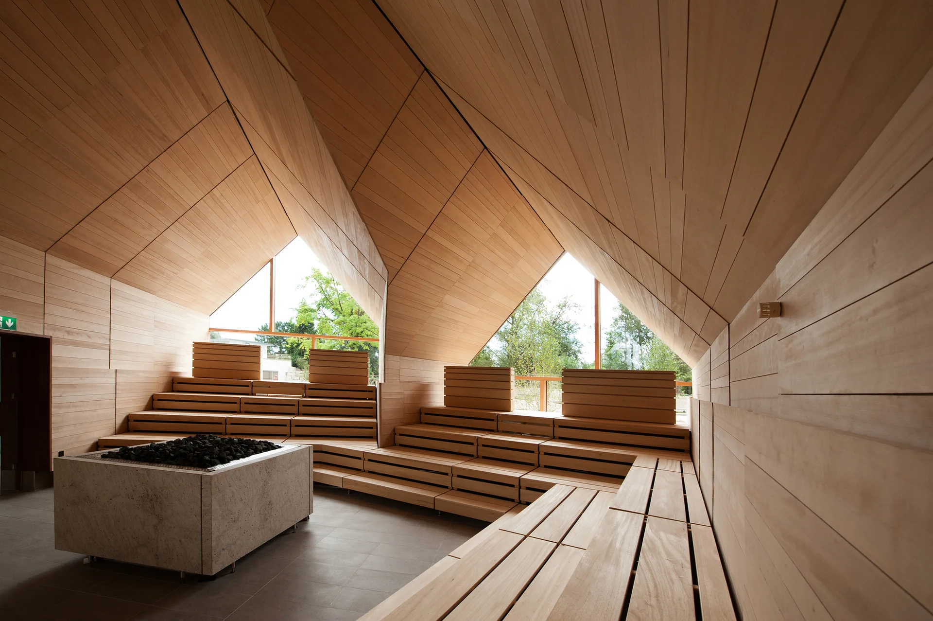 A modern wood-paneled sauna features tiered bench seating, a central stone-filled heater, and triangular windows under an angular vaulted ceiling.