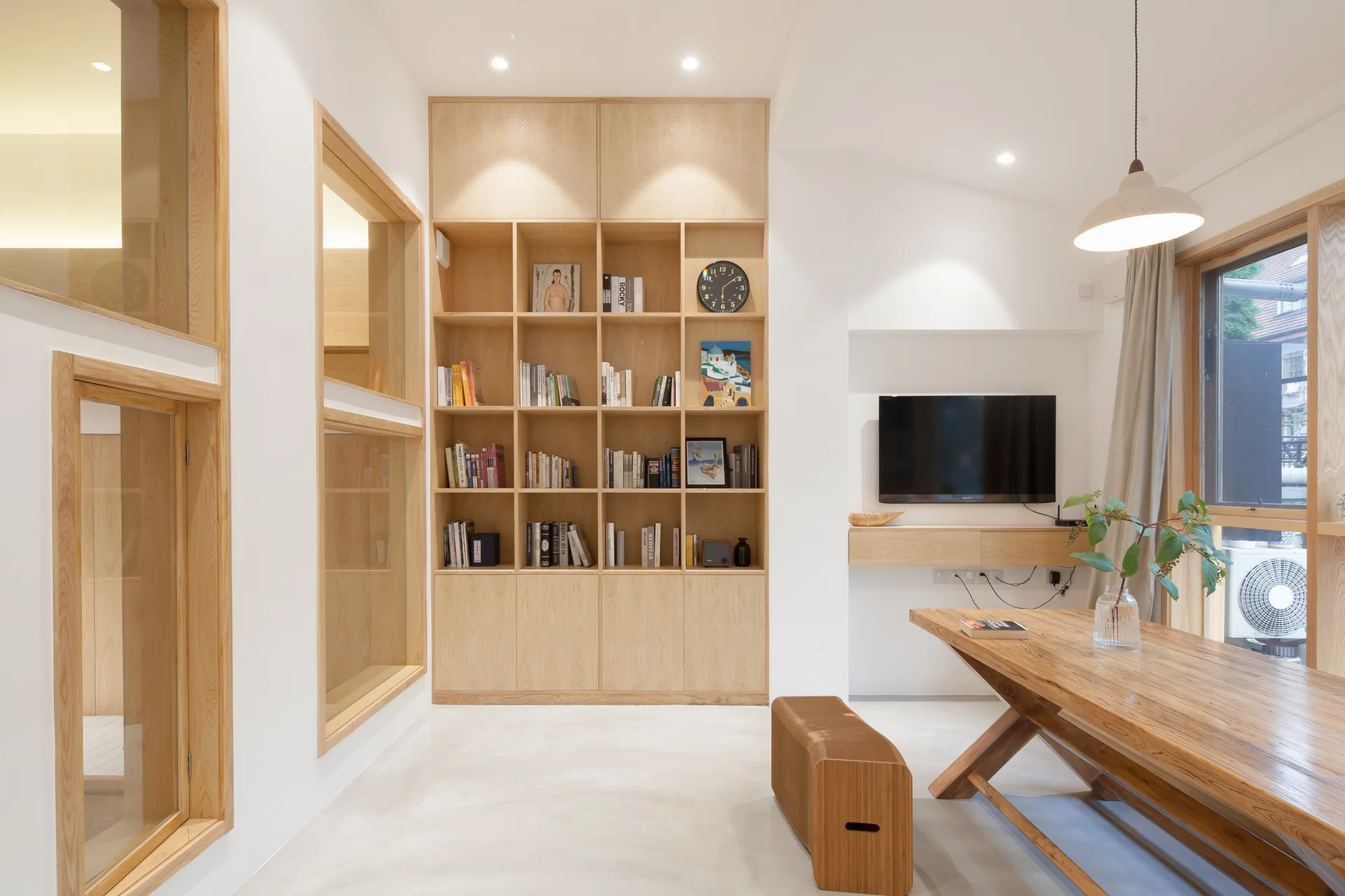 A minimalist dining room features a large light wood table, a tall built-in bookshelf, and internal wood-framed windows set against white walls.
