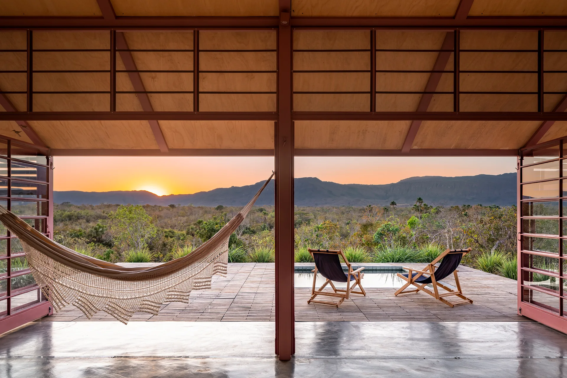 A covered wood deck features a woven hammock, two folding lounge chairs, and a small pool overlooking a sunset view of distant hills.