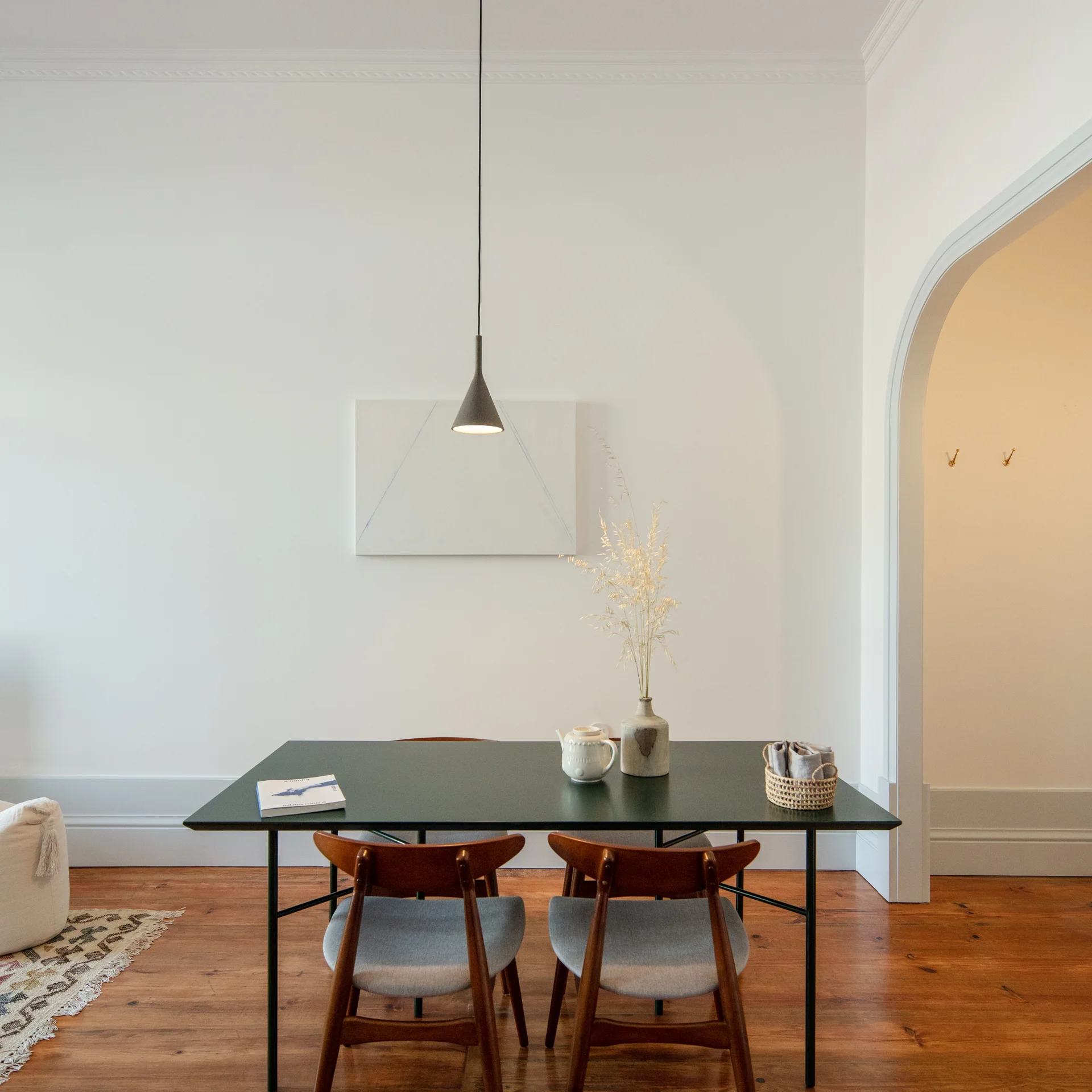 A dark green dining table paired with wooden mid-century chairs stands on hardwood floors beneath a single slim cone pendant light.