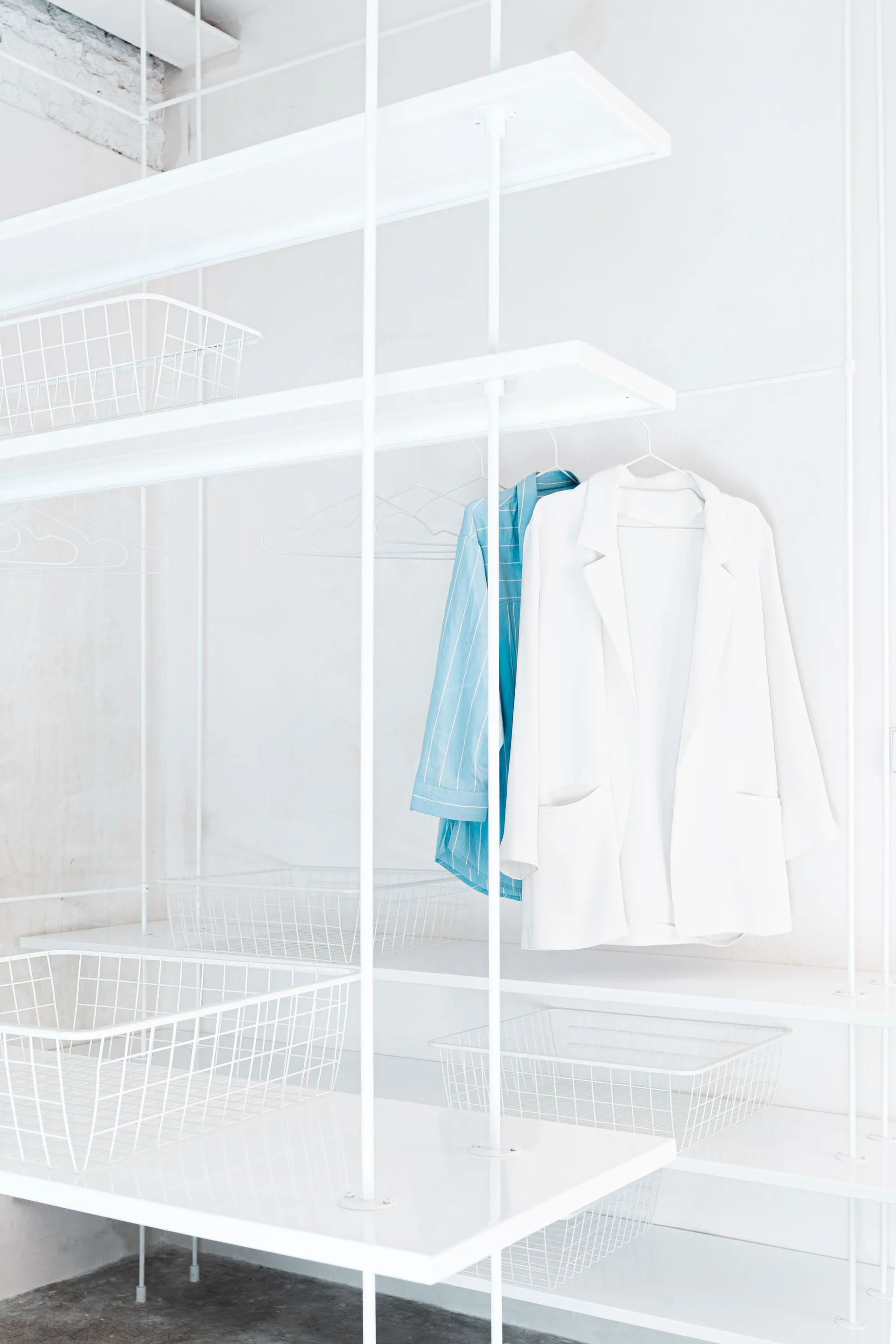 A minimalist white metal wire closet shelving system holds matching wire storage baskets and hanging garments against a white wall.