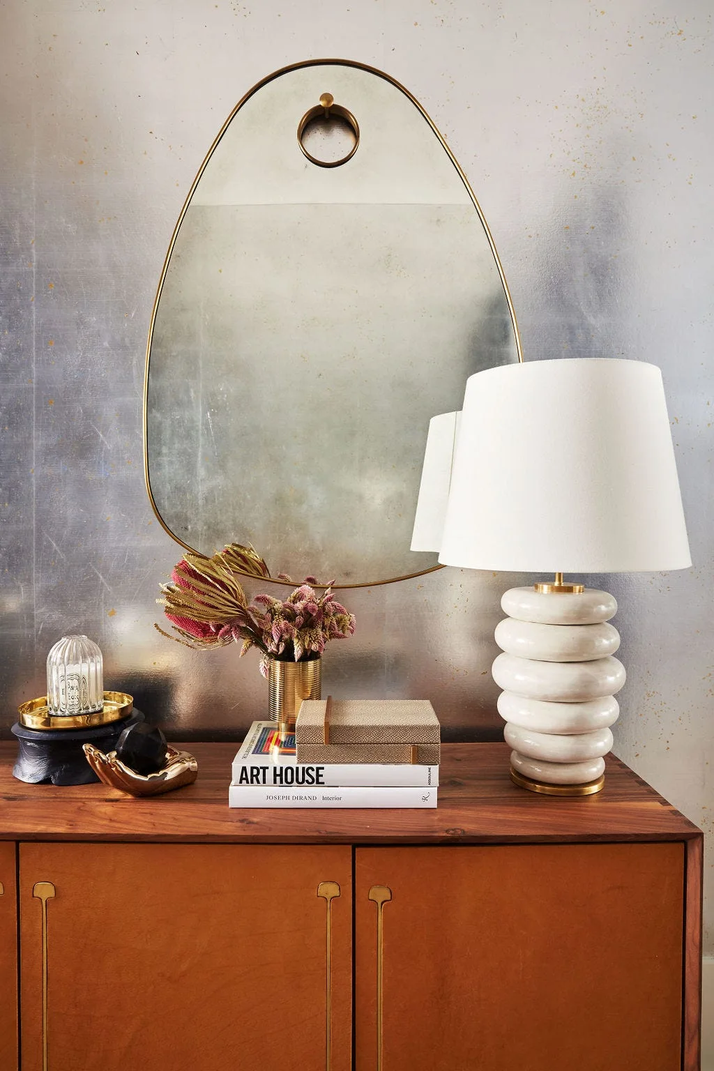 An oval brass mirror hangs above a wood and leather console table styled with a ribbed white table lamp, books, and decorative brass objects.