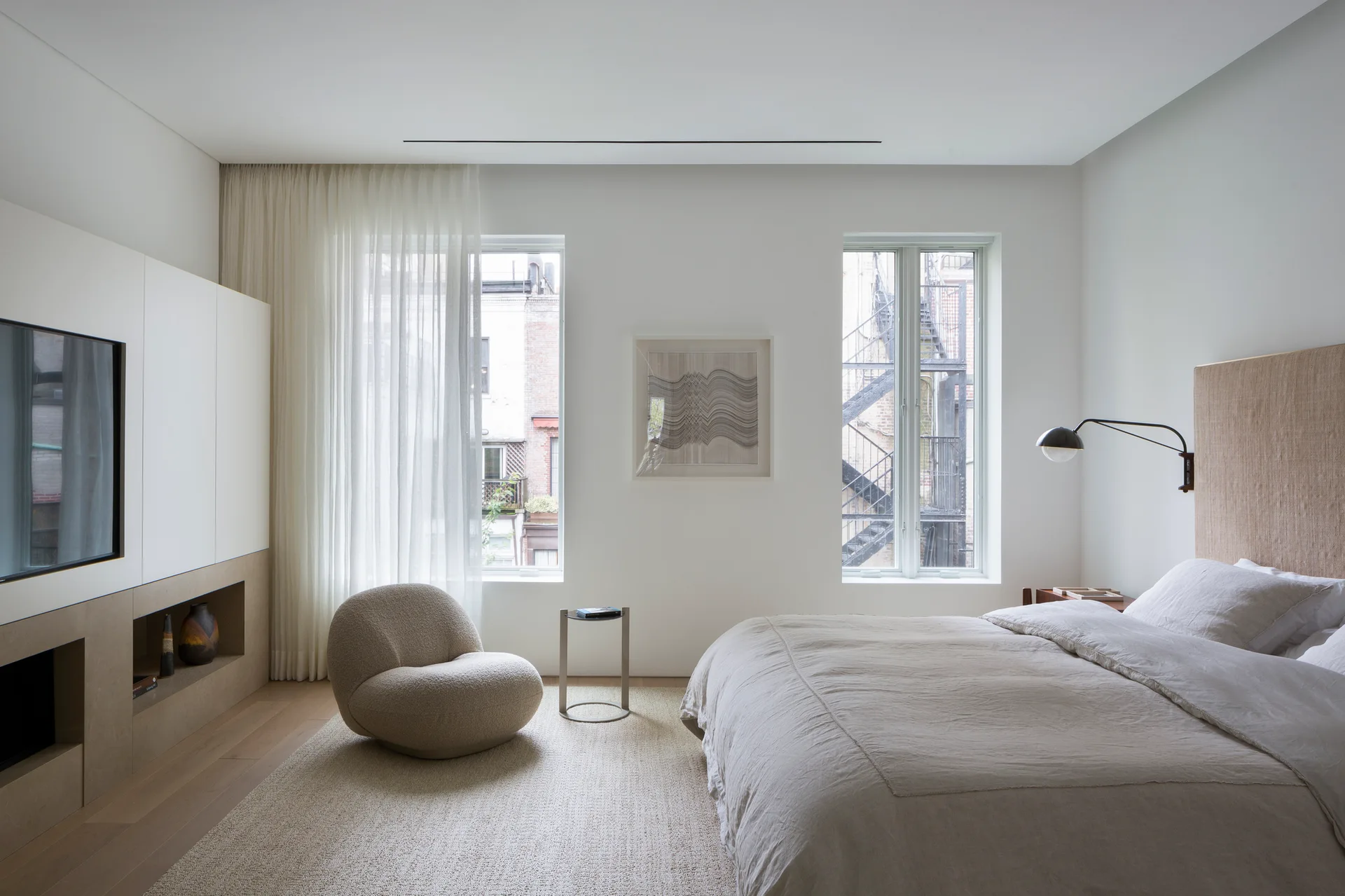 A modern bedroom features a linen-covered bed, a round boucle lounge chair, sheer white curtains, and a custom white media wall unit.