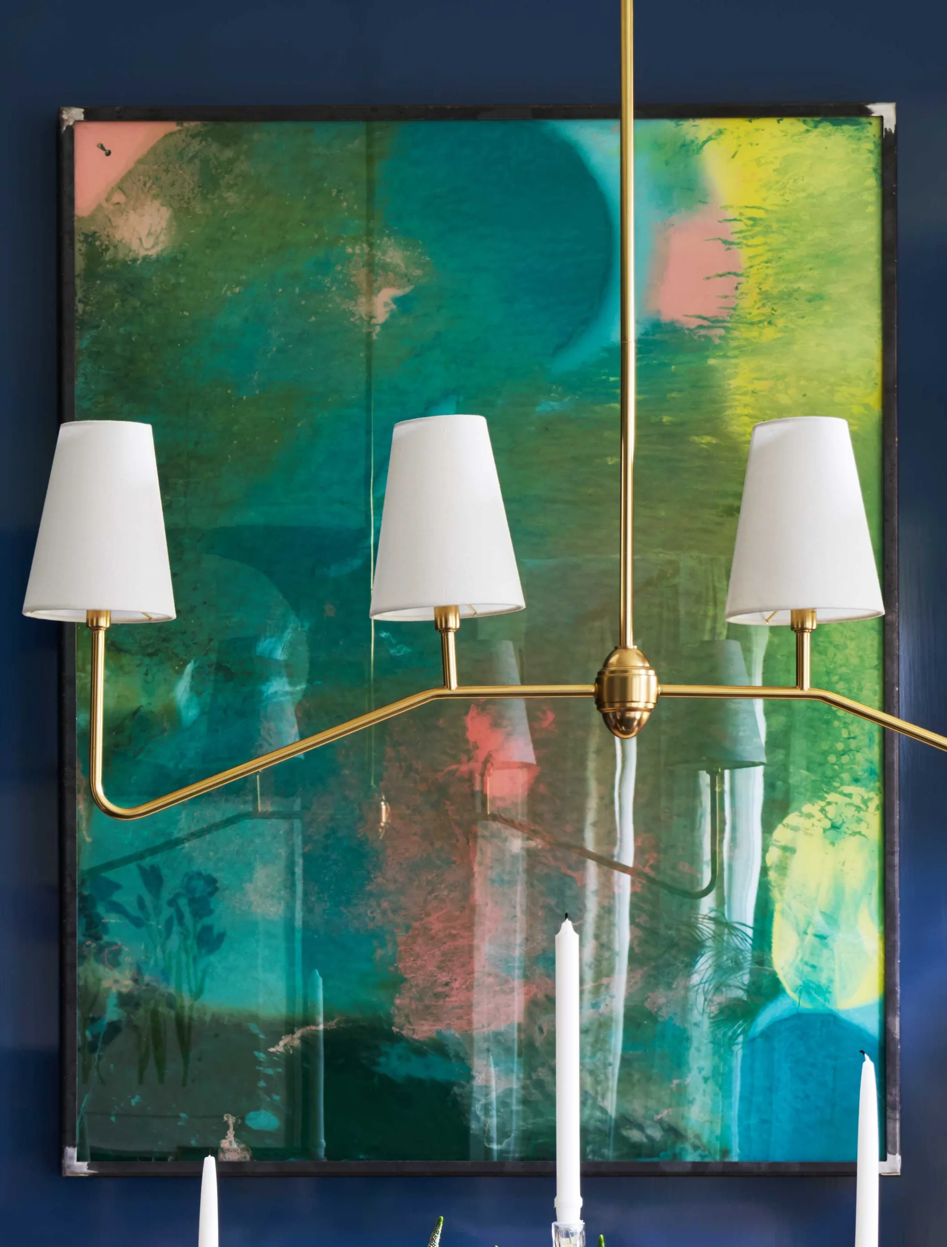 A brass multi-arm chandelier with white fabric shades hangs in front of a large, colorful abstract painting on a deep navy wall.