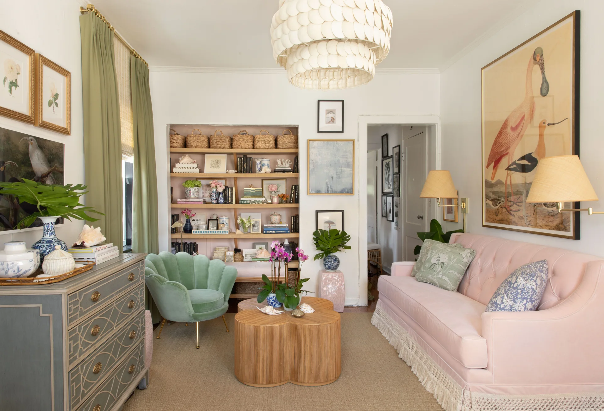 An eclectic living room featuring a pink tufted sofa with fringe trim, a green scalloped armchair, a ribbed wood coffee table, and built-in shelving.