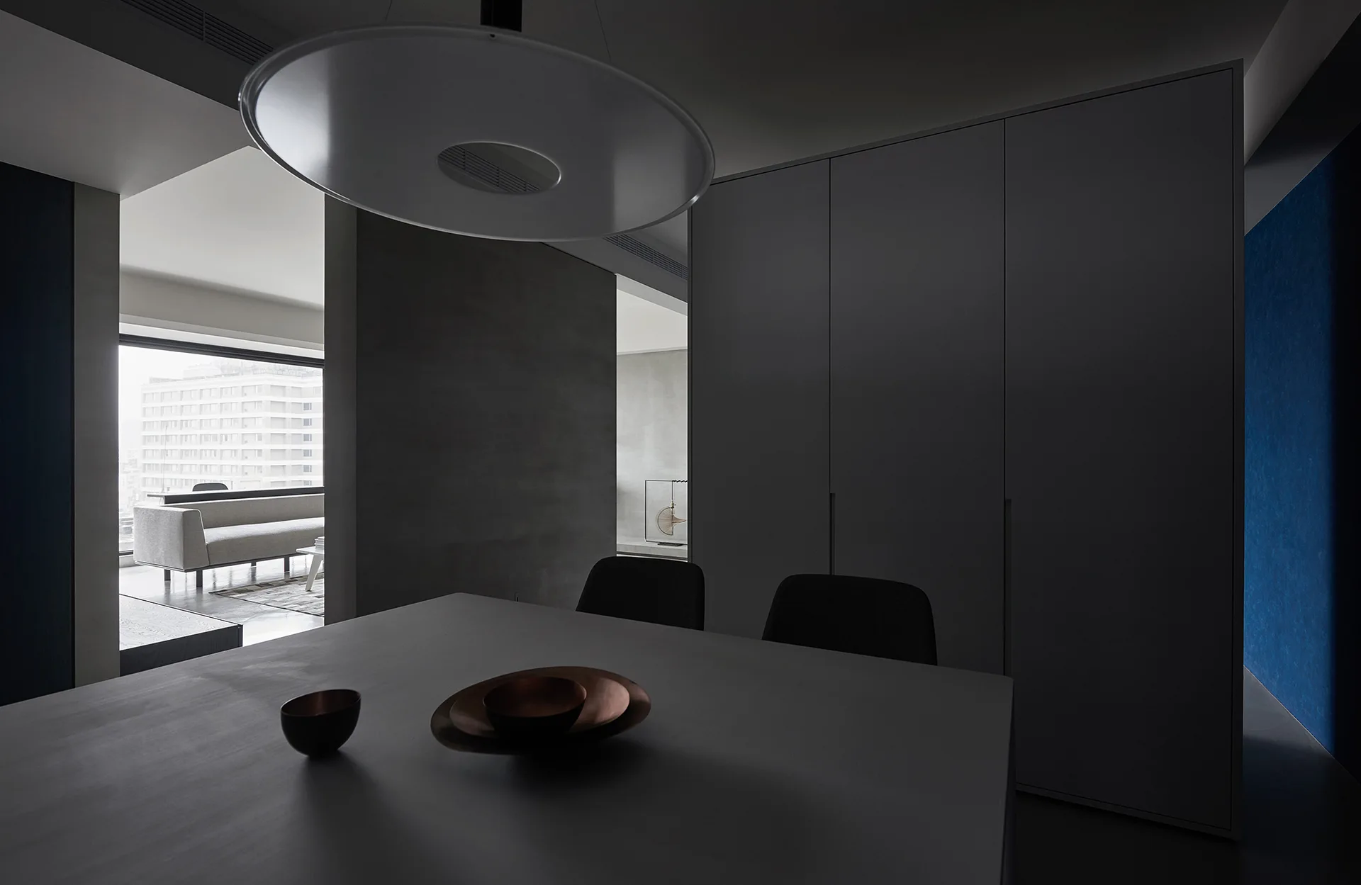 A dark-toned dining room features a large disc pendant light above a grey table with decorative bowls next to a tall grey cabinet wall.