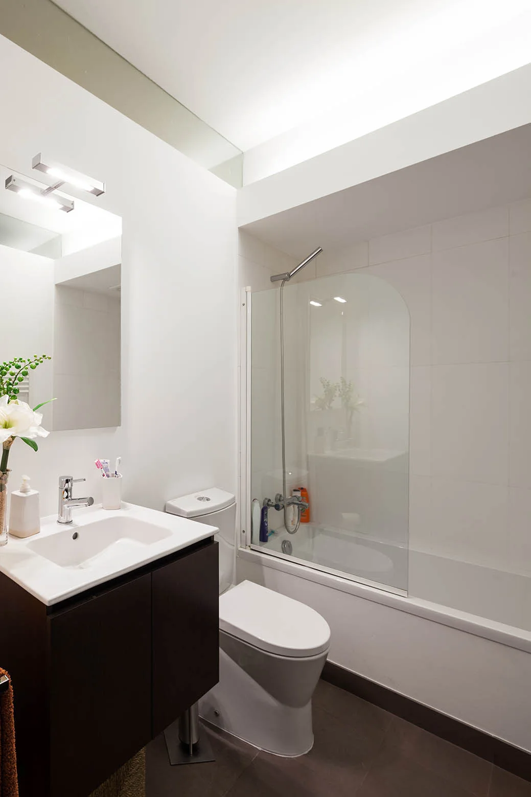 A compact modern bathroom features a dark wood floating vanity, a white toilet, and a bathtub with a curved glass shower screen.