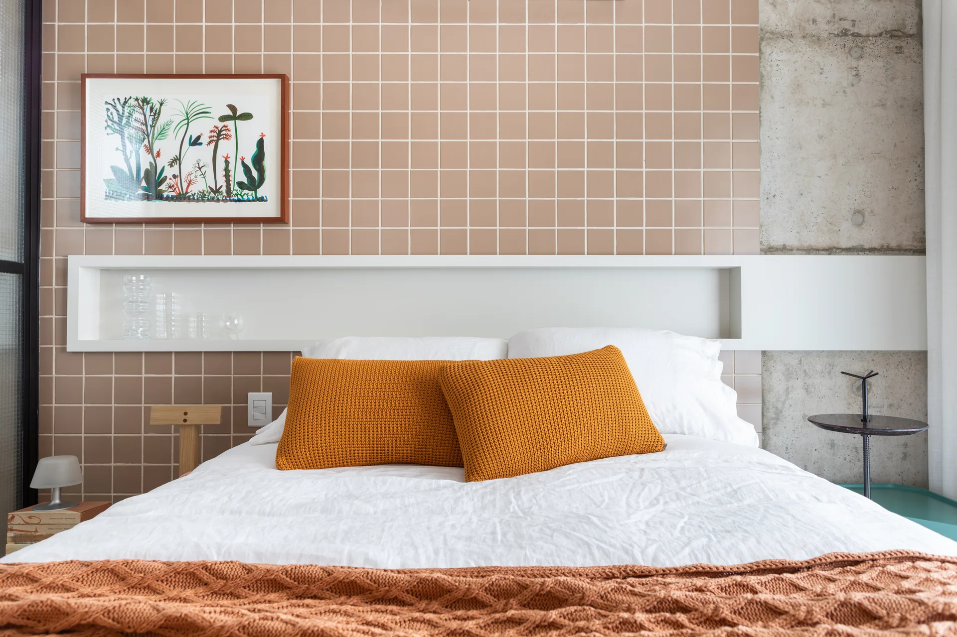 A bed with white bedding and orange pillows sits against a beige grid-tiled wall featuring a built-in horizontal white niche shelf.
