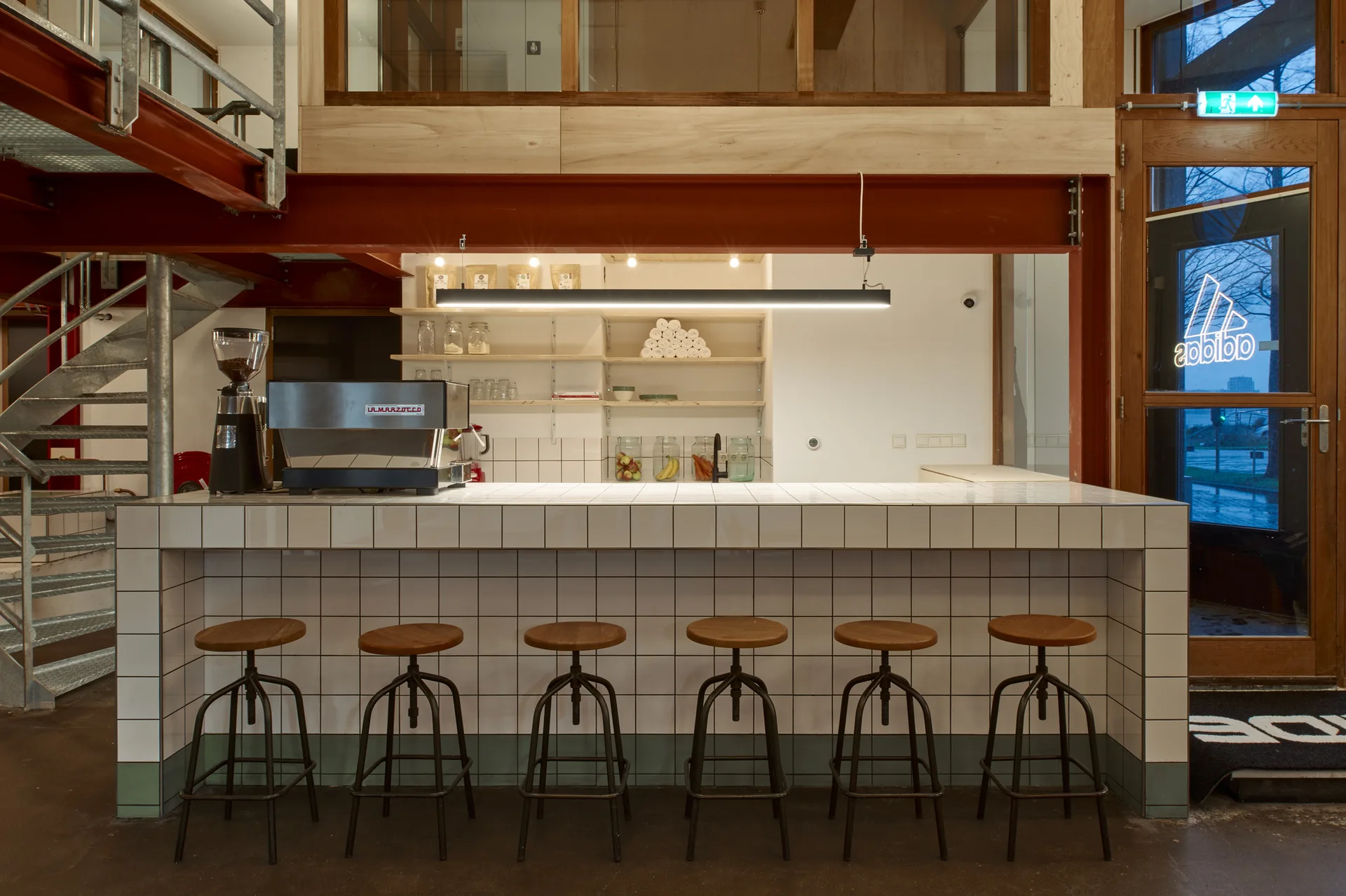 An industrial coffee bar features a white-tiled counter lined with six wooden stools beneath an exposed red steel beam.