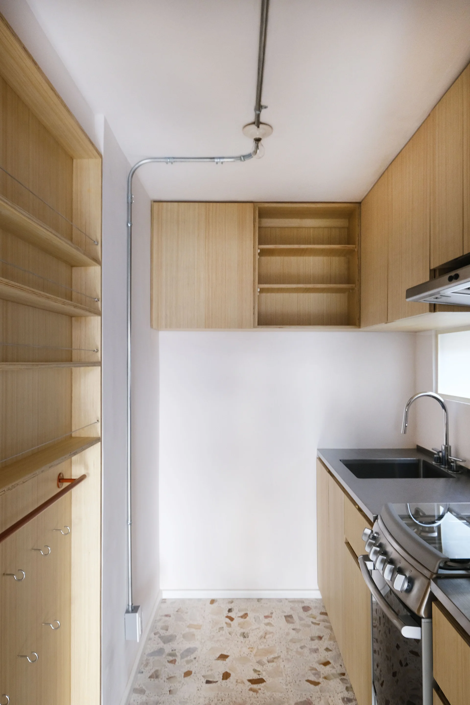 A compact modern kitchen featuring blonde wood cabinets, a speckled terrazzo tiled floor, and an exposed metal wall conduit.