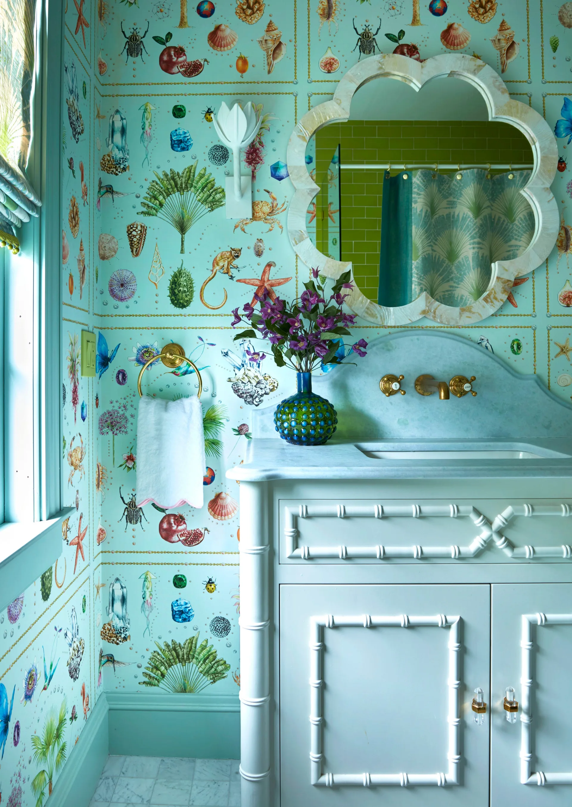 A powder room features light blue natural-science patterned wallpaper behind a white faux-bamboo vanity, a scalloped mirror, and gold fixtures.