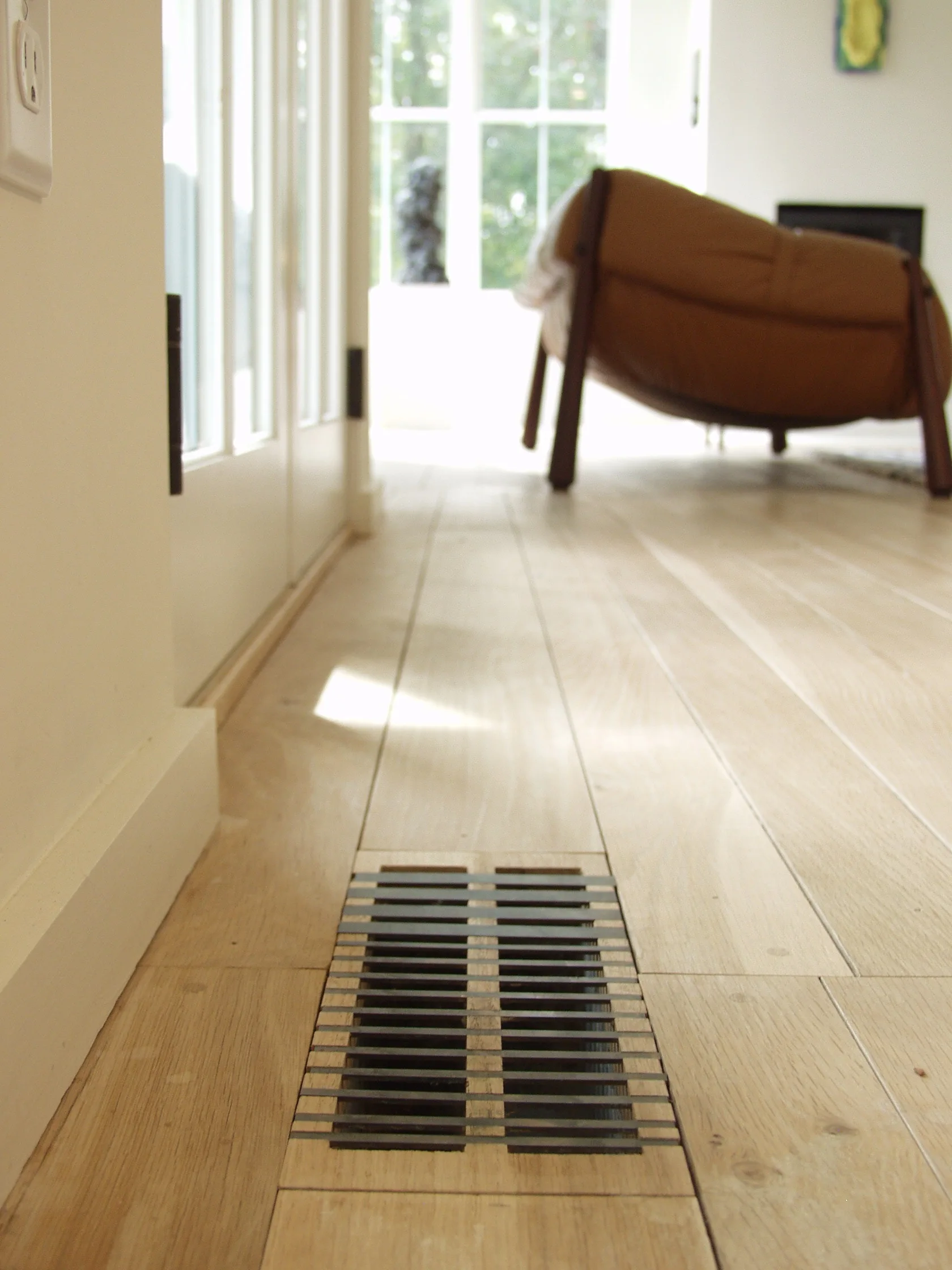 A metal floor vent integrated into light hardwood planks, with white walls and a brown leather armchair in the background.