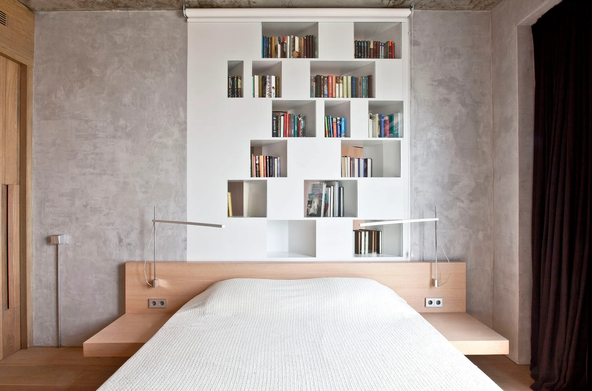 A light wood platform bed rests against a concrete wall integrated with a white geometric cutout shelving unit.