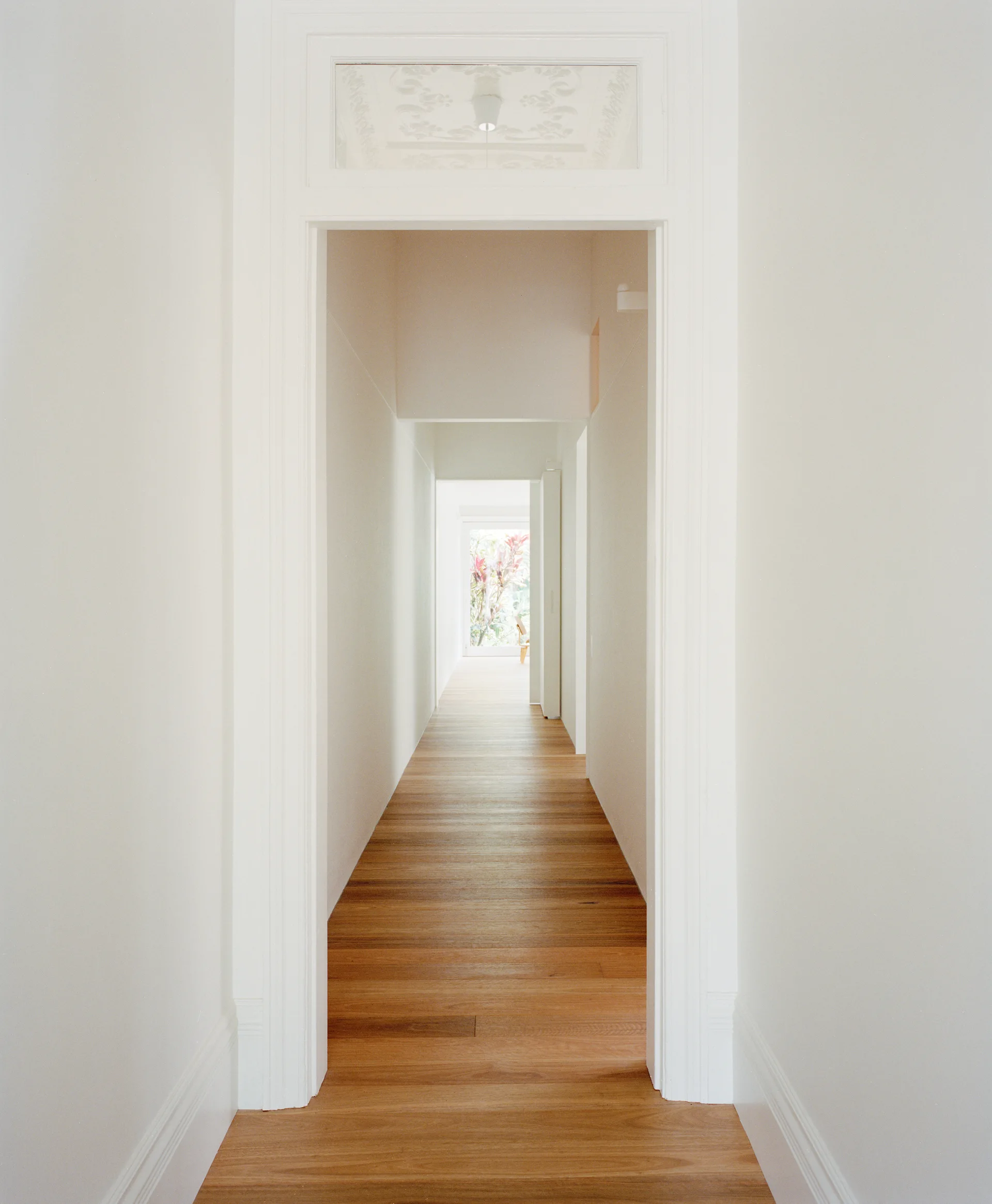 A long white residential hallway features natural hardwood plank floors and a decorative glass transom window above a prominent door frame.