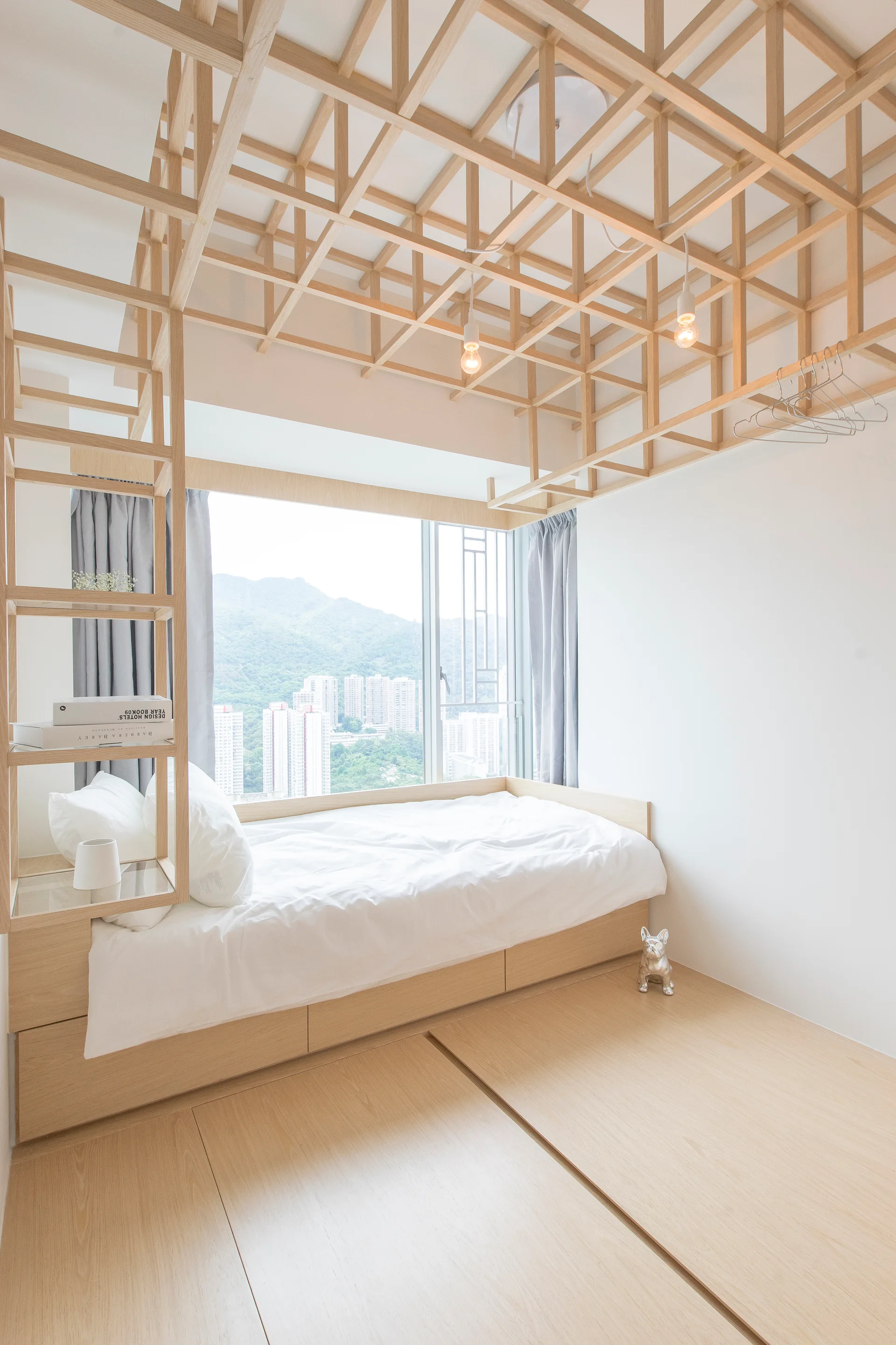 A compact modern bedroom features a light wood platform bed beneath a matching wooden ceiling lattice structure next to a large window.