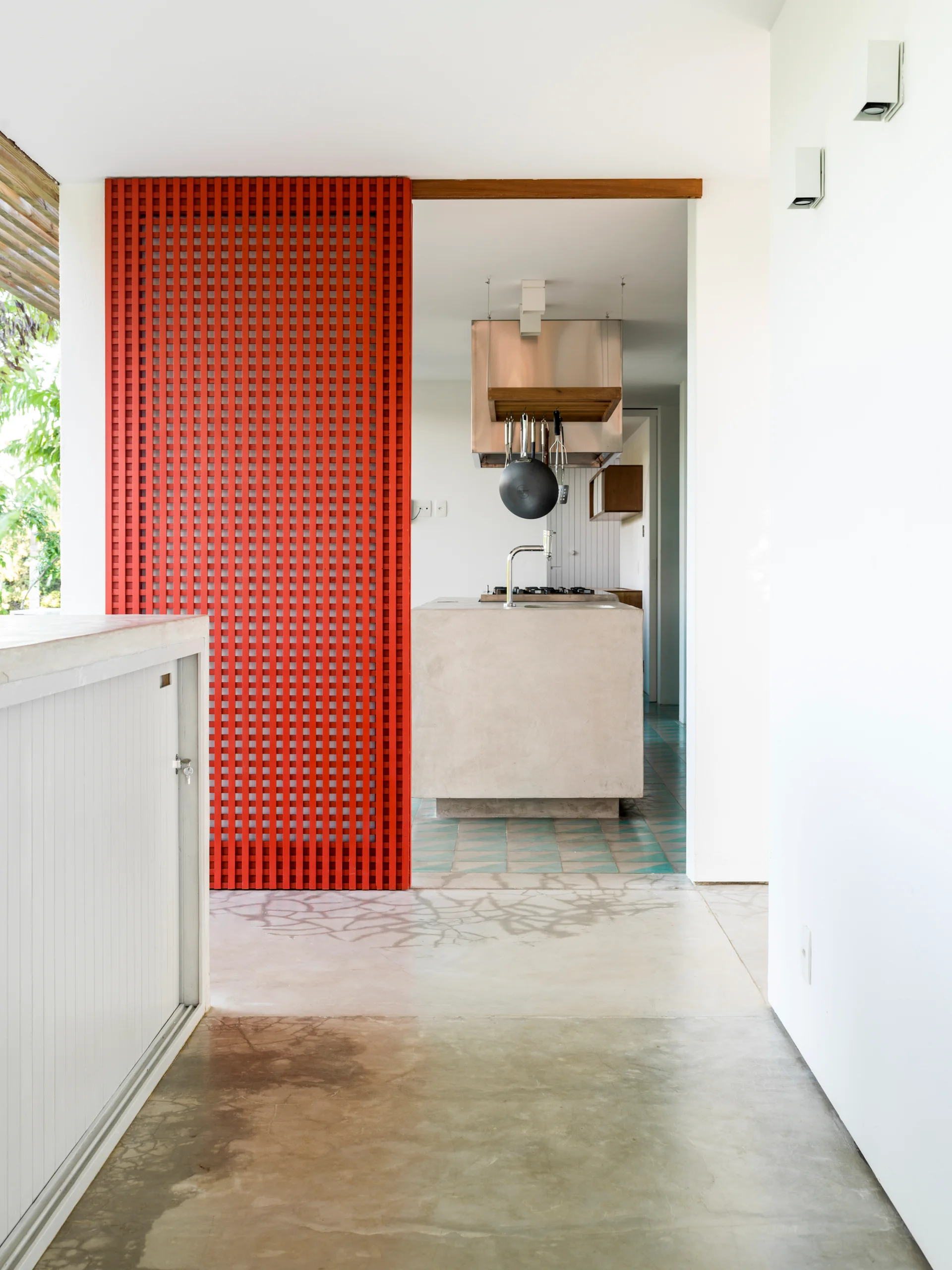 A view through a concrete corridor toward a kitchen featuring a concrete island and a bright red grid-patterned sliding partition screen.