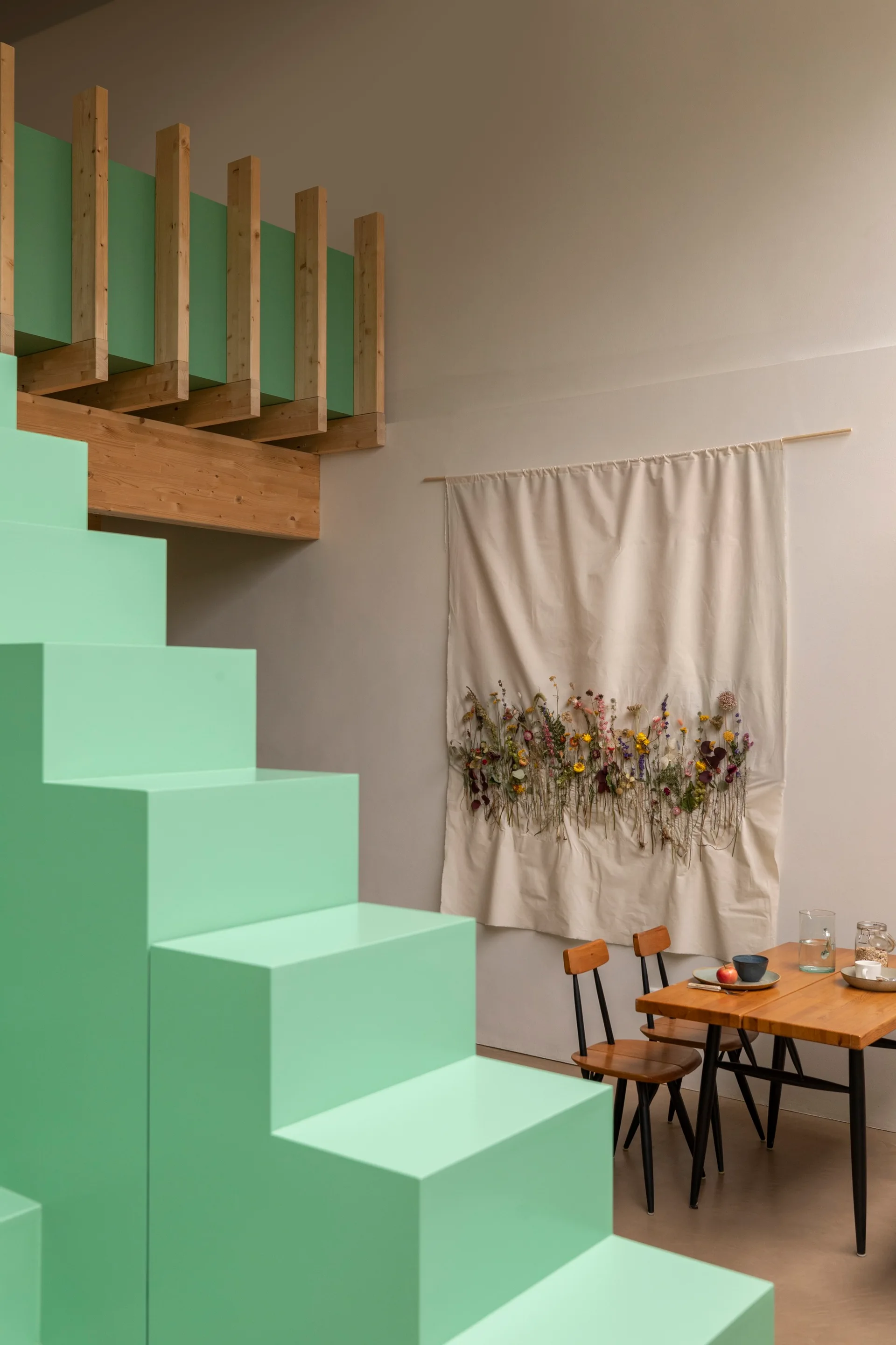 A dining area features a bright mint green geometric staircase, a wooden table and chairs, and a canvas wall hanging adorned with dried flowers.