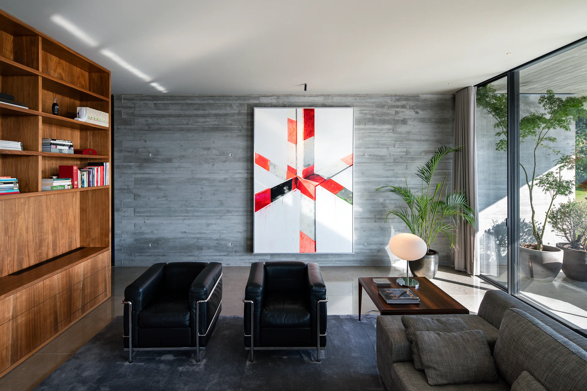 A minimalist living room featuring a textured concrete wall, a large wooden bookcase, two black leather armchairs, and a large abstract painting.