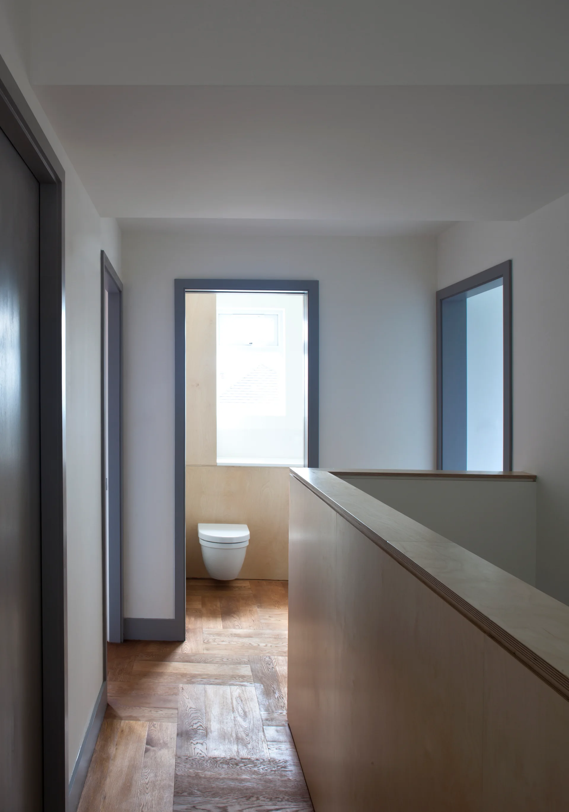 A minimalist hallway landing with herringbone wood floors, white walls, gray door trim, and a plywood-clad staircase railing.