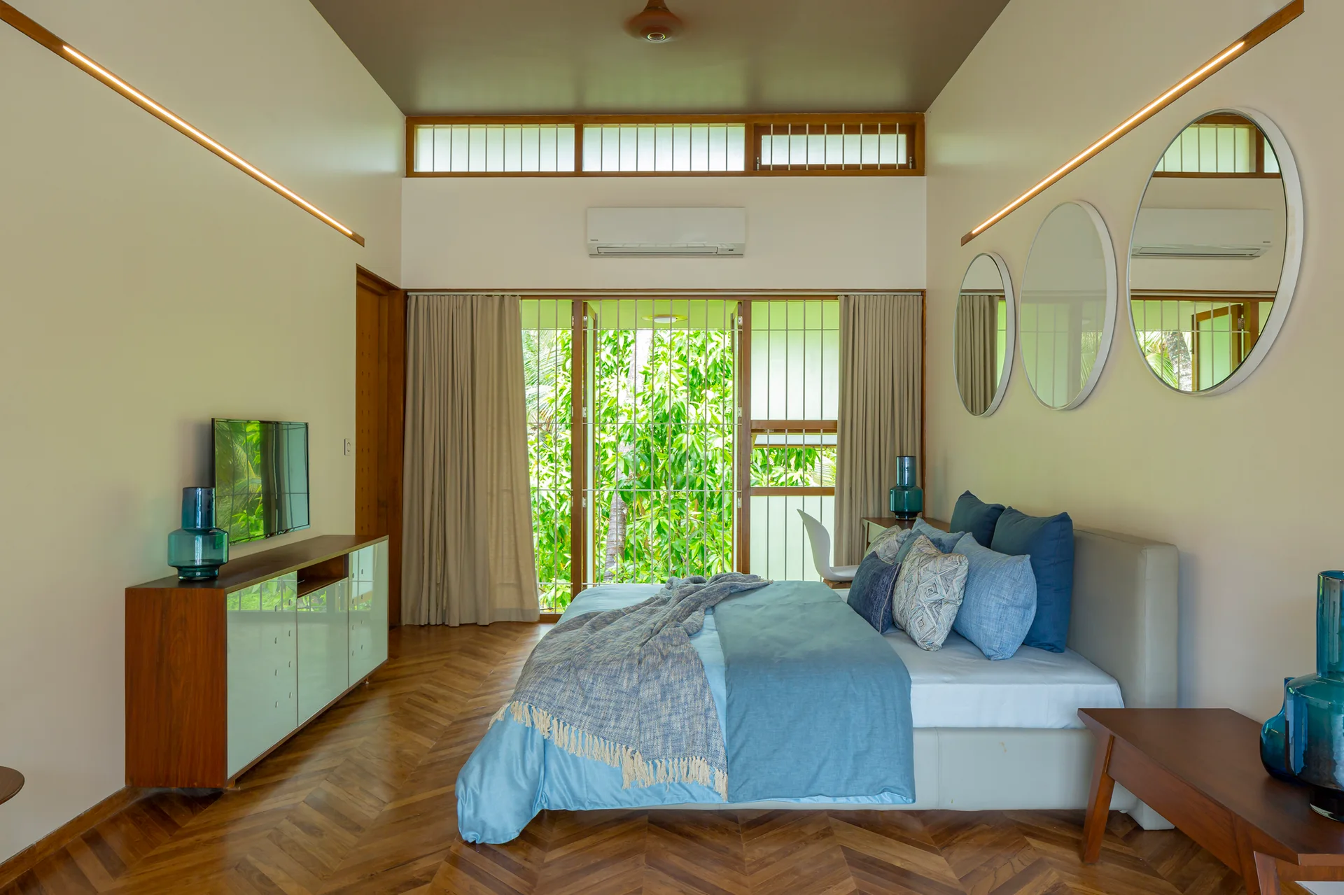 A contemporary bedroom featuring herringbone wood floors, a bed with blue sheets, a wall mounted television, and three round wall mirrors.