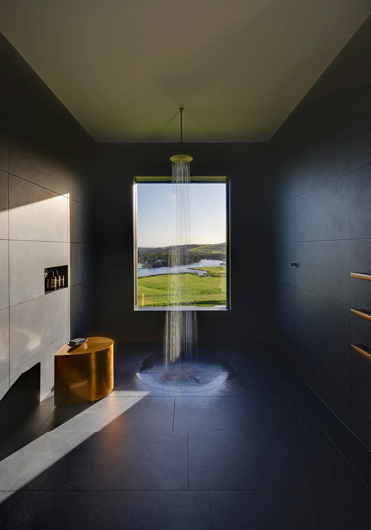 A dark charcoal tiled walk-in shower features a gold curved stool, a ceiling-mounted brass showerhead, and a large window looking out at a grassy river valley.