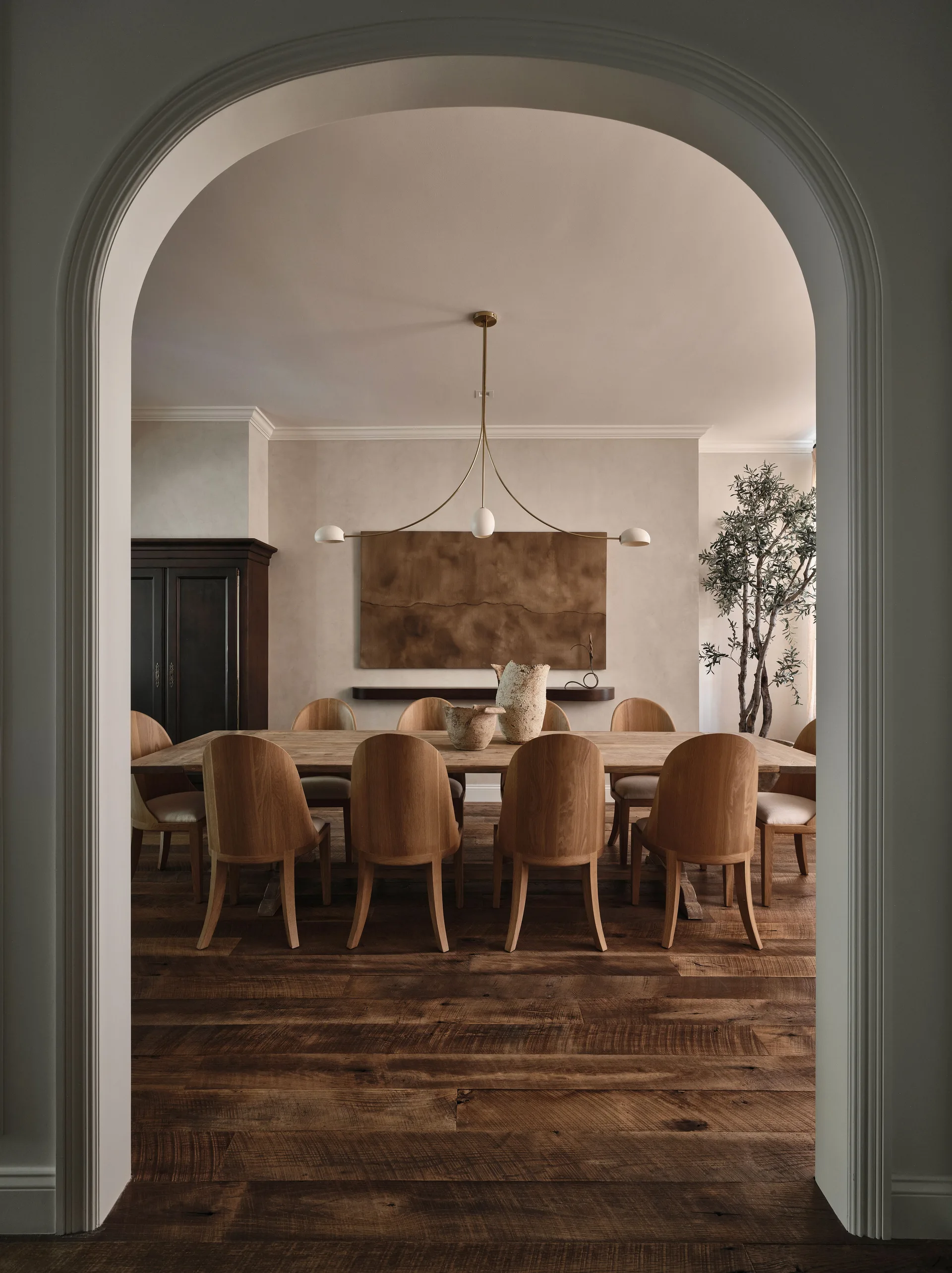 A dining room framed by a white archway features a long wooden table with curved chairs, a modern brass chandelier, and neutral beige walls.
