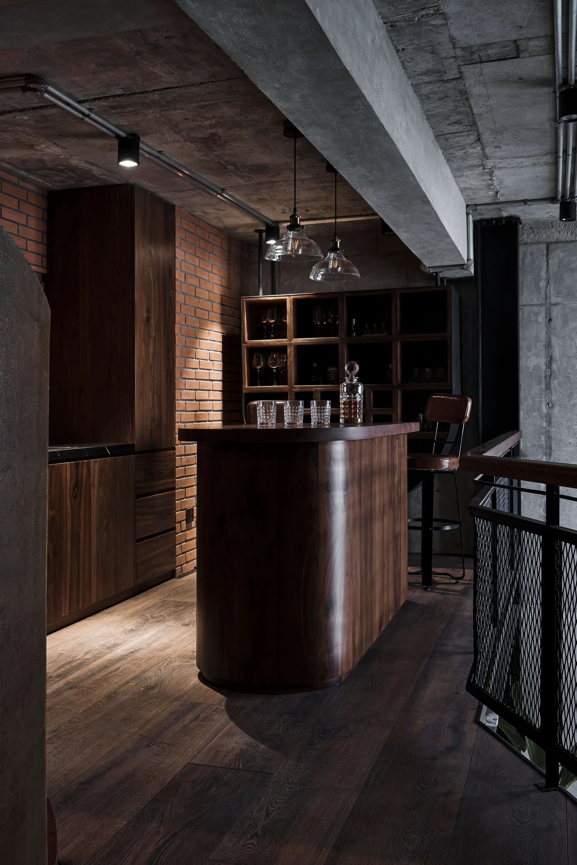 An industrial home bar featuring a curved dark wood counter, exposed concrete ceiling, a brick wall, and glass pendant lights.