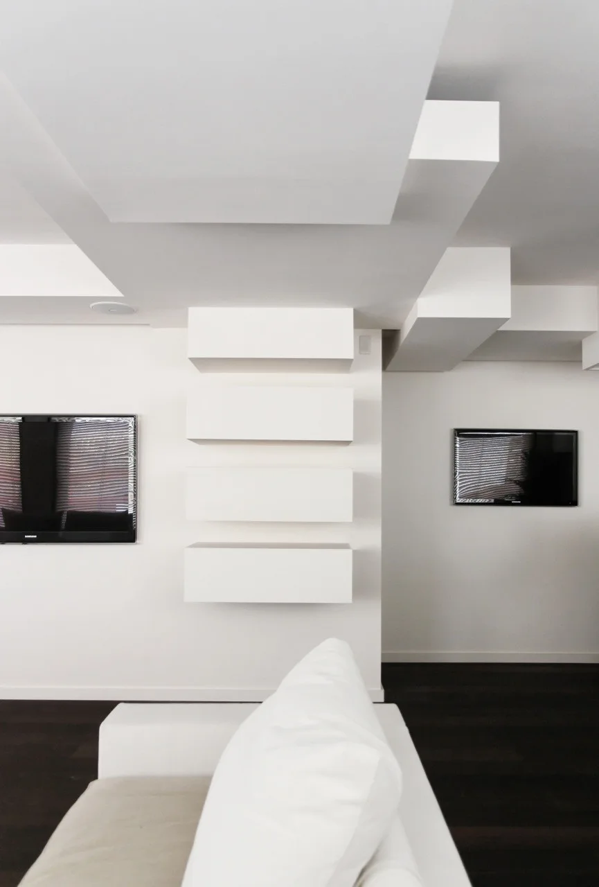 A minimalist white living room features a white sofa, dark wood floors, and stacked geometric floating shelves beneath a dropped ceiling.