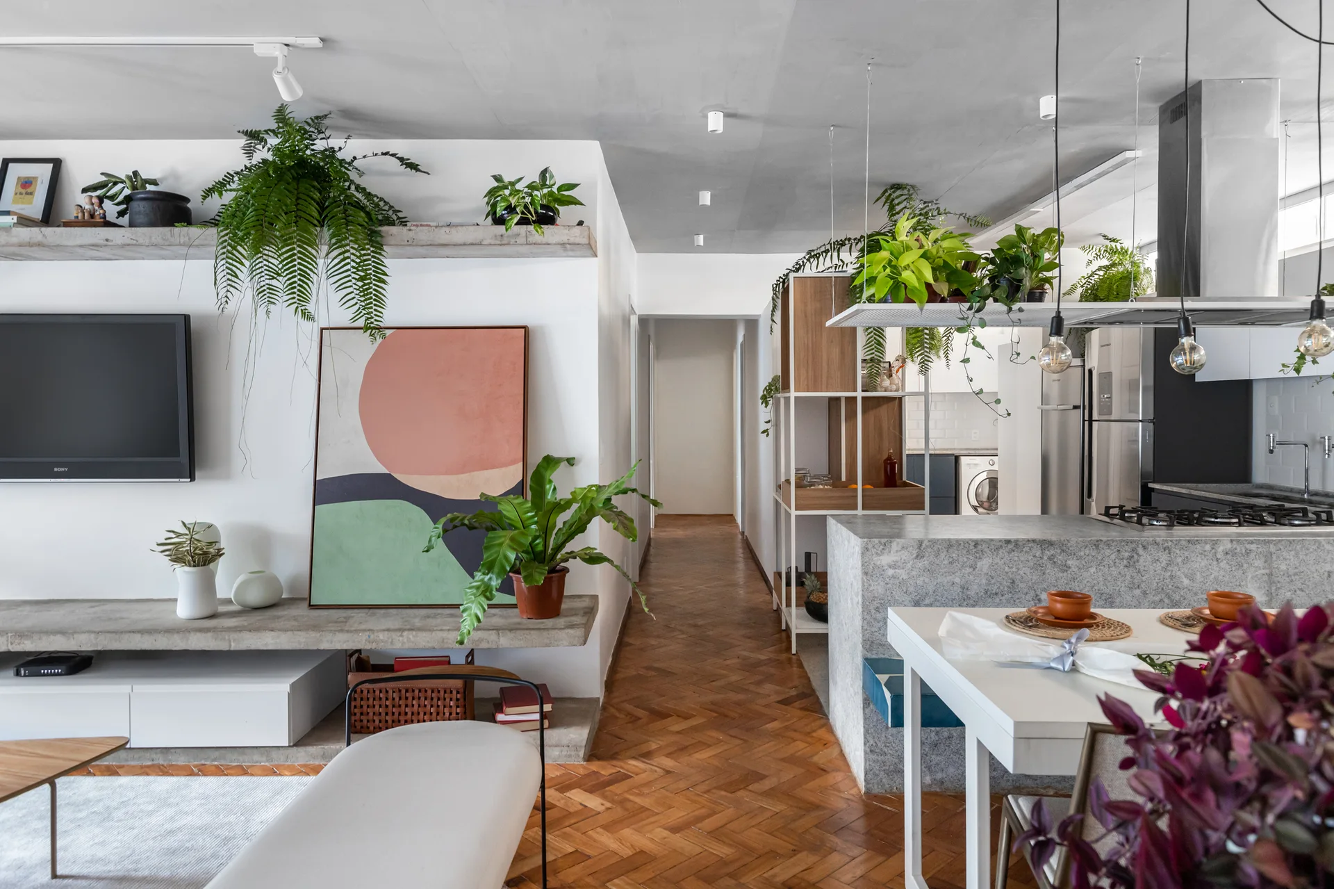 An open-plan living room and kitchen featuring herringbone parquet wood flooring, floating concrete shelves, and a stone-look kitchen island.