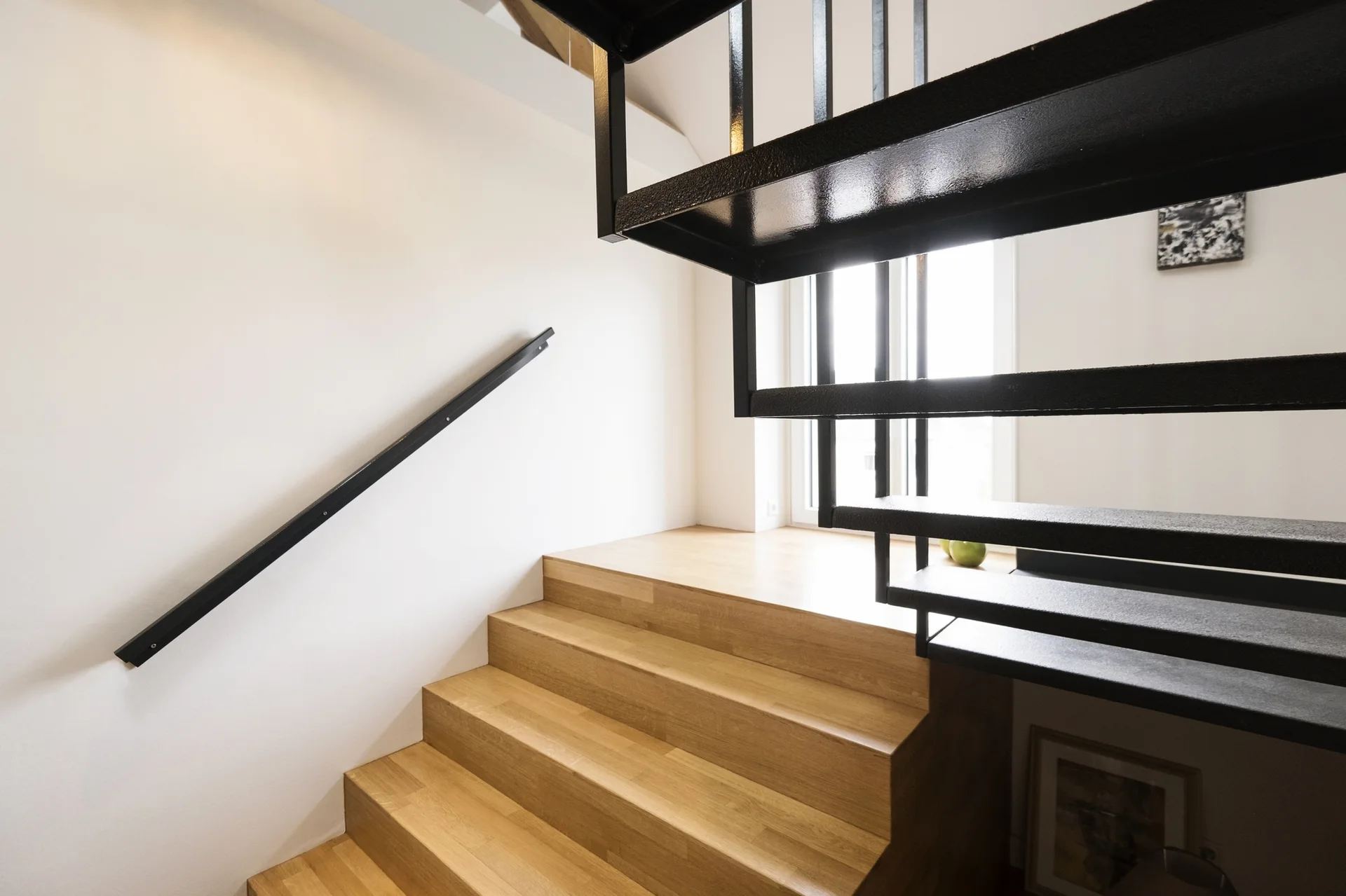 A modern staircase featuring light wood steps on the lower half and open-riser black metal stairs above, set against white walls with a slim black handrail.