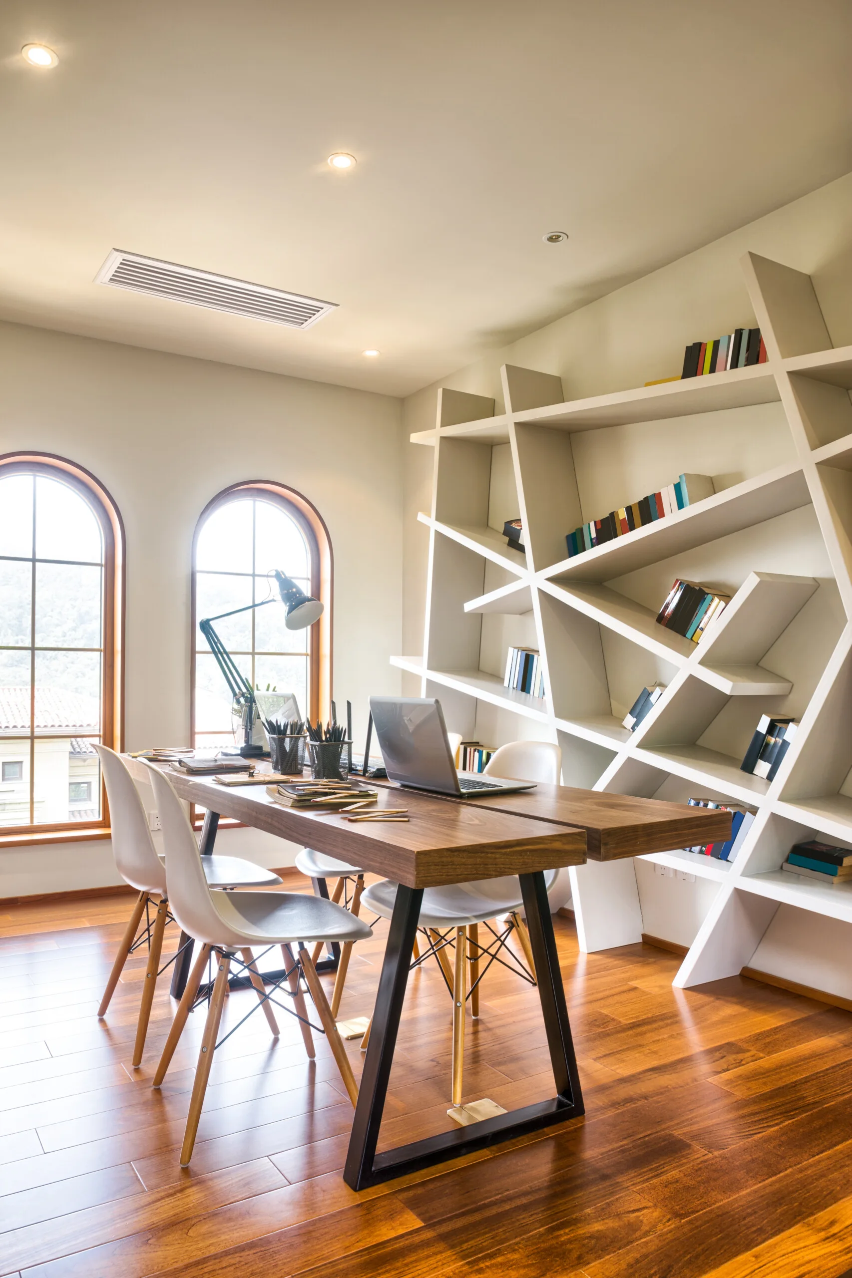 A home office features a wooden desk with white chairs, two arched windows, and a large white geometric shelving unit filled with books.