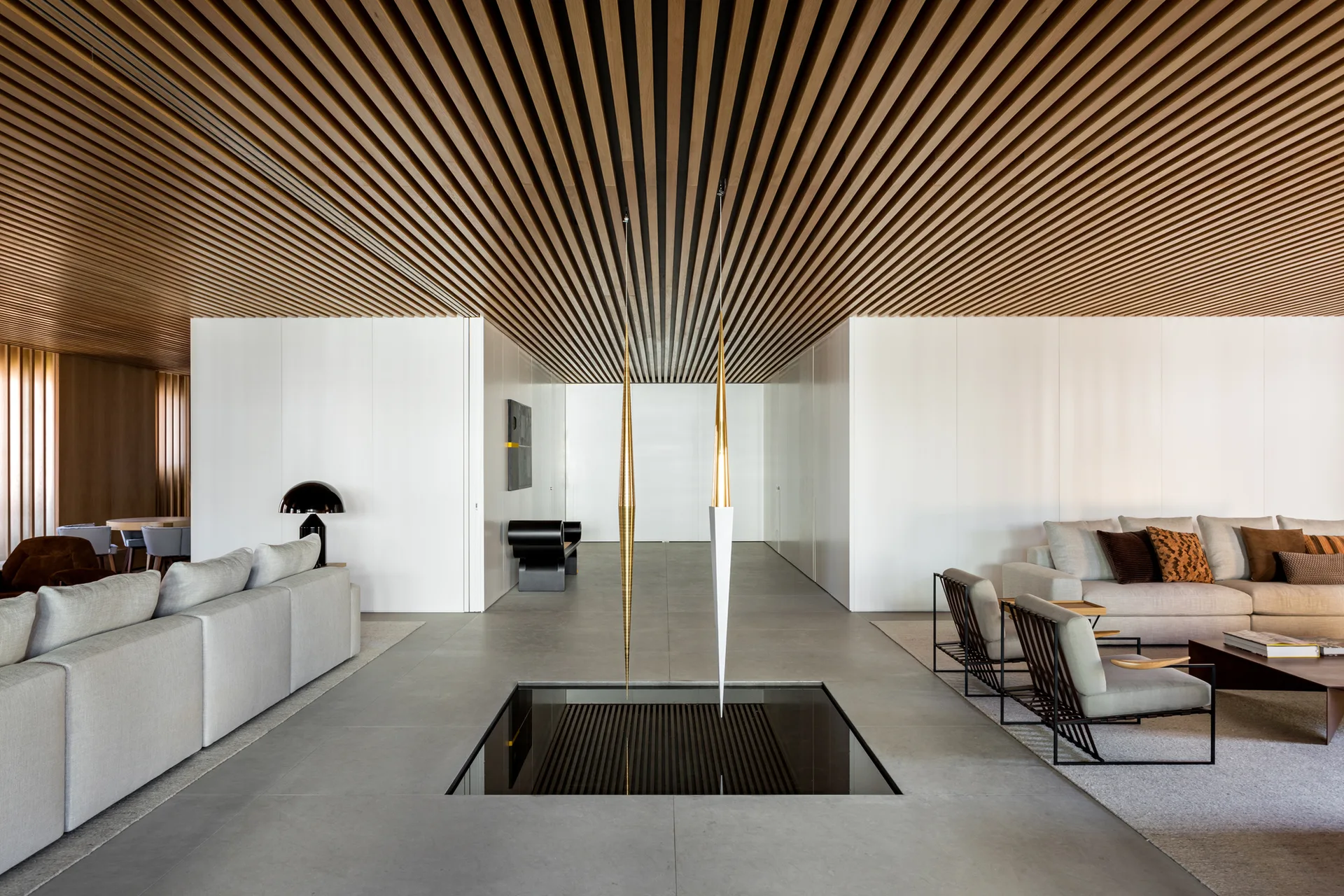 A modern living room features a linear slatted wood ceiling, a square recessed water feature in the stone floor, grey sofas, and white walls.