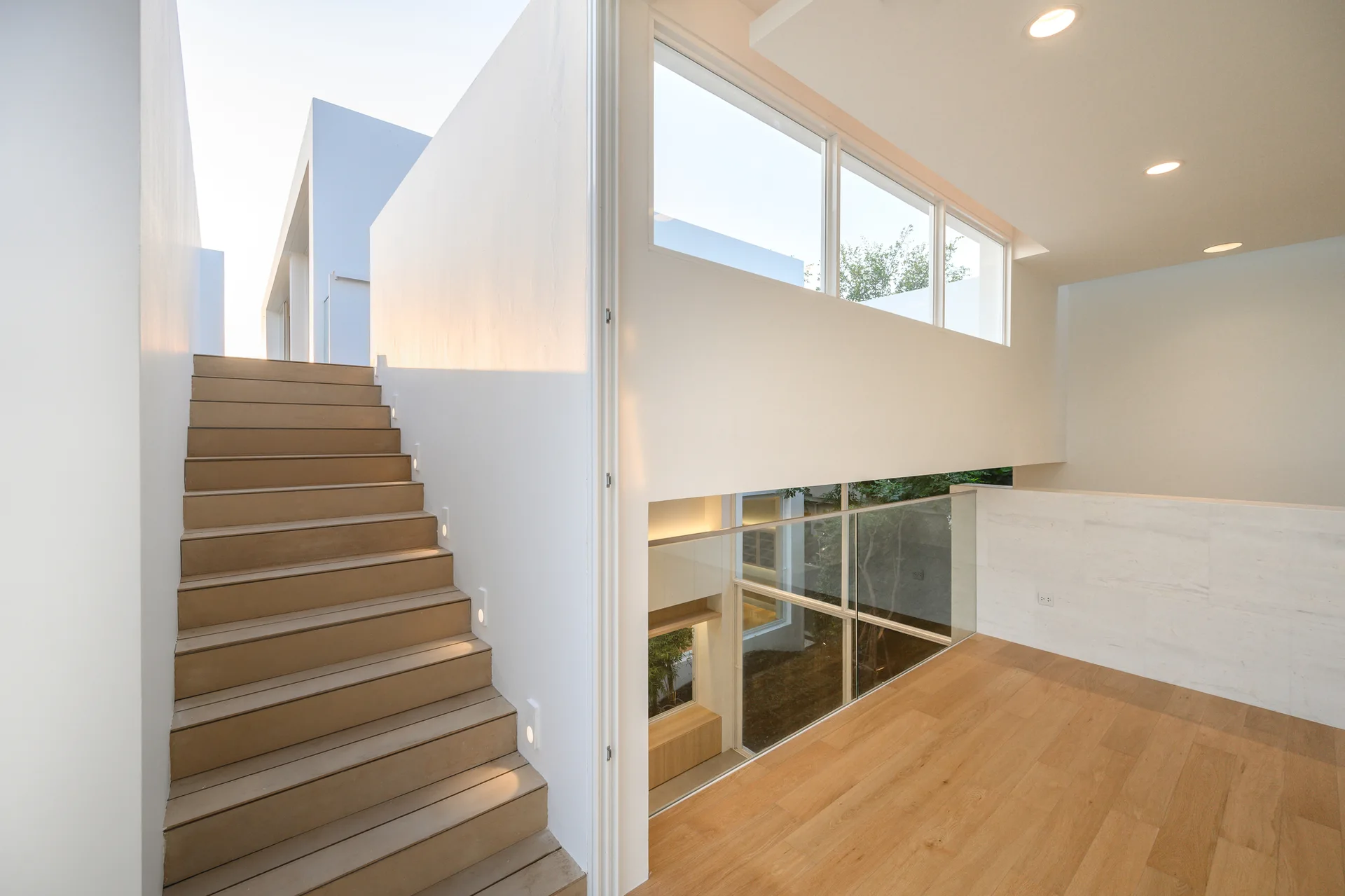 A modern wood staircase with step lights stands next to a light wood landing featuring a glass railing and horizontal windows under a white ceiling.