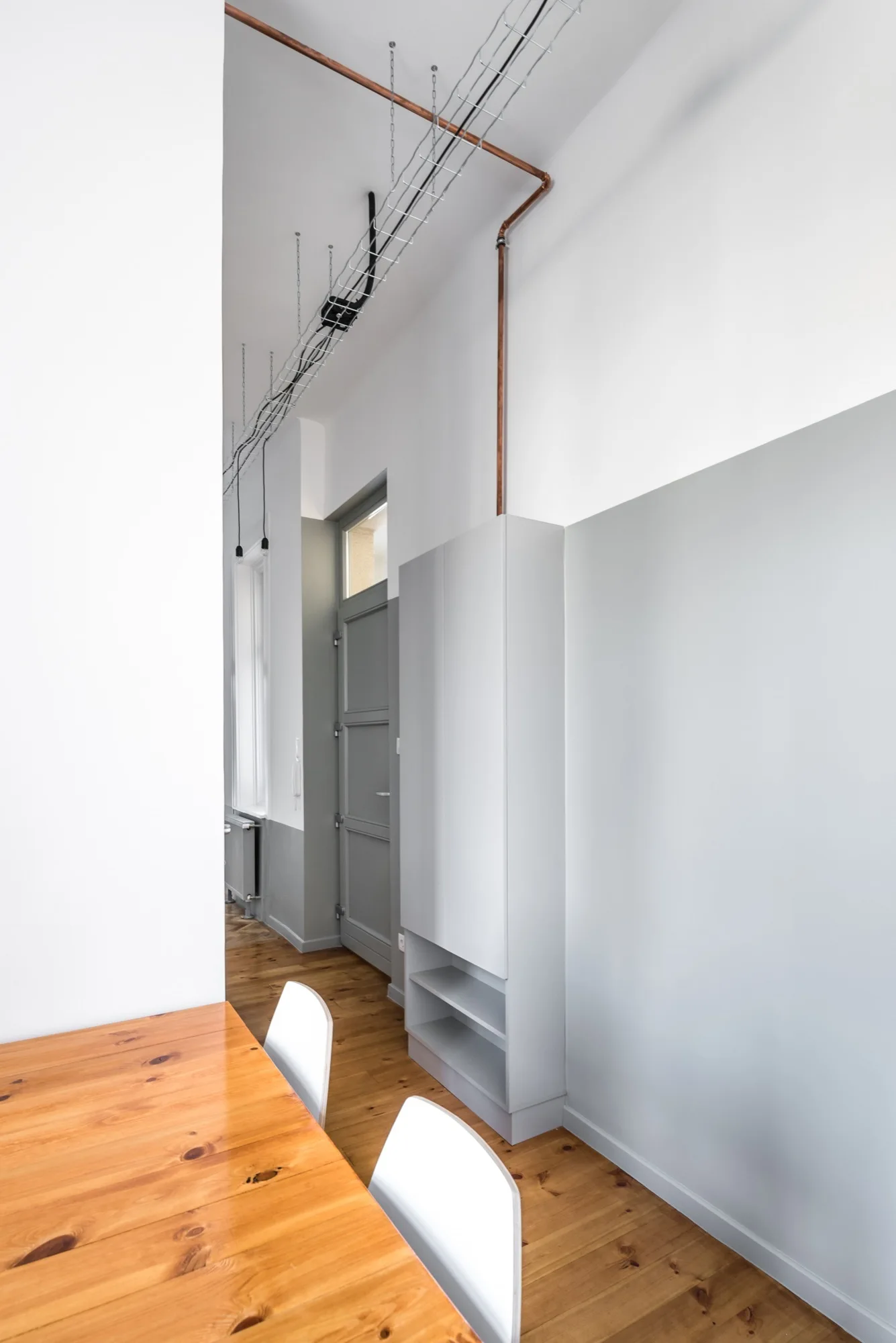 A minimalist hallway features a tall gray storage cabinet against a gray accent wall under exposed copper piping and a metal ceiling cable tray.