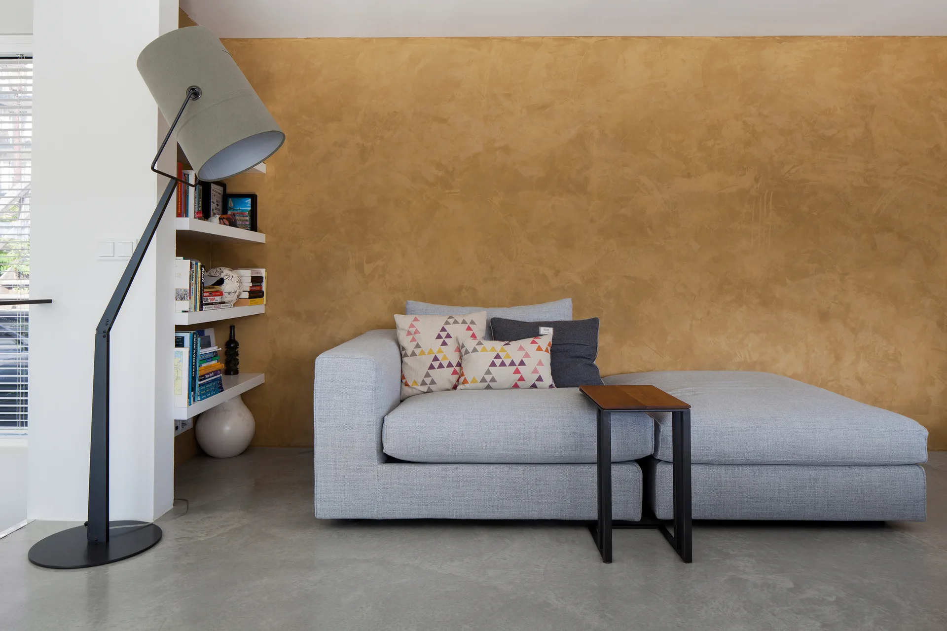 A light gray sofa stands against a textured mustard gold wall next to white built in shelves and a large gray floor lamp.