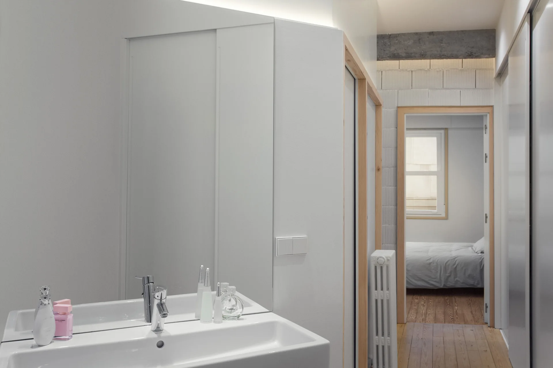 A minimalist white bathroom sink and large frameless mirror looking toward a doorway with light wood trim that opens into a bedroom.