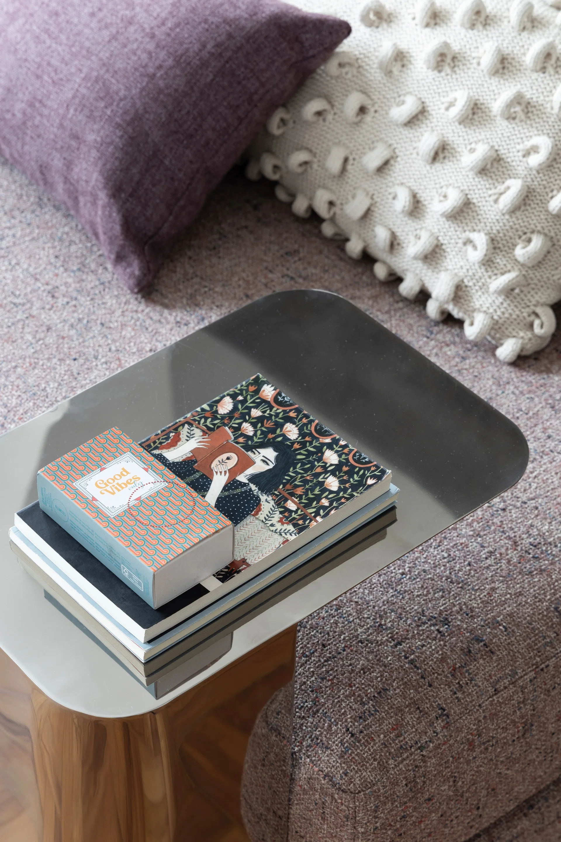 A reflective metal side table holds a stack of books next to a fabric sofa decorated with purple and cream bobble knit pillows.