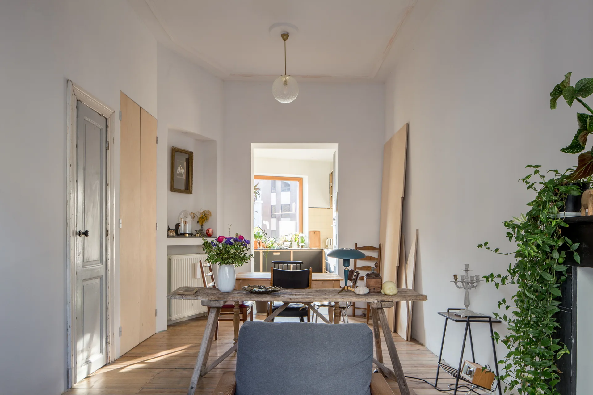 An eclectic dining room features a weathered wood trestle table, mixed dining chairs, and an open doorway leading to a bright kitchen.