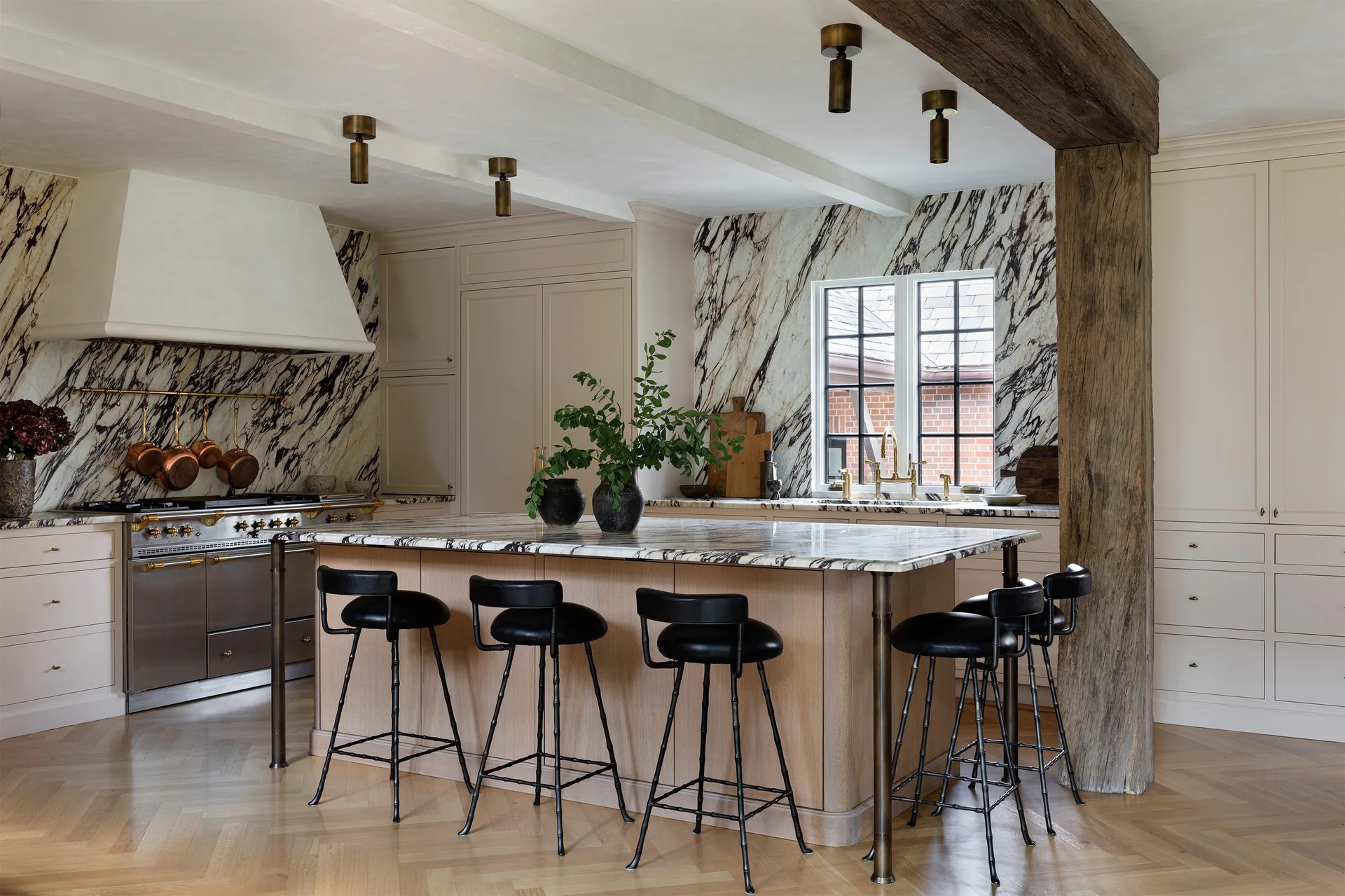 A cream kitchen features a marble-topped island with black stools, heavily veined marble backsplashes, and a prominent rustic wooden ceiling beam.
