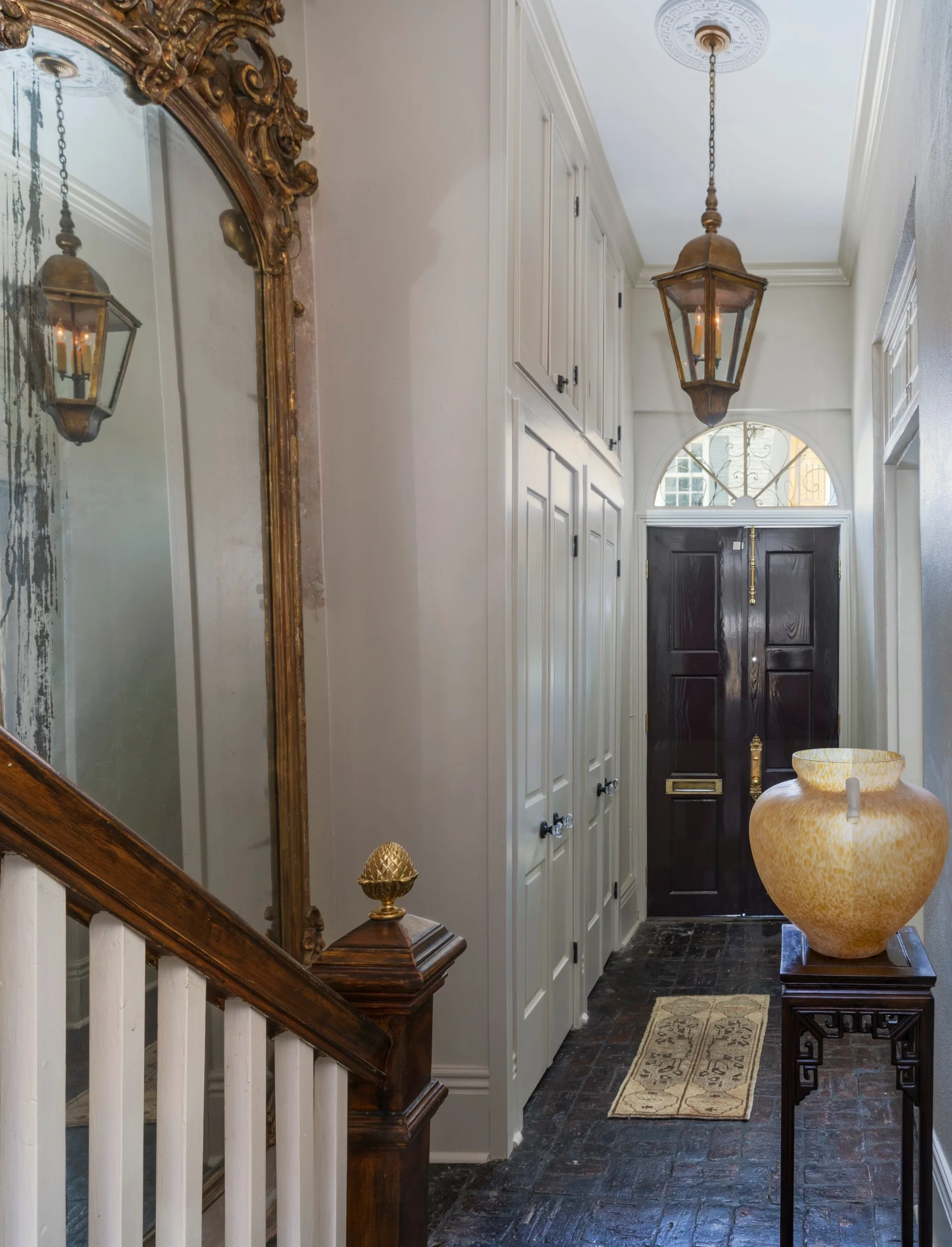 A narrow traditional entryway features a dark double door, built-in cream cabinets, an ornate gold-framed mirror, and a hanging lantern light.
