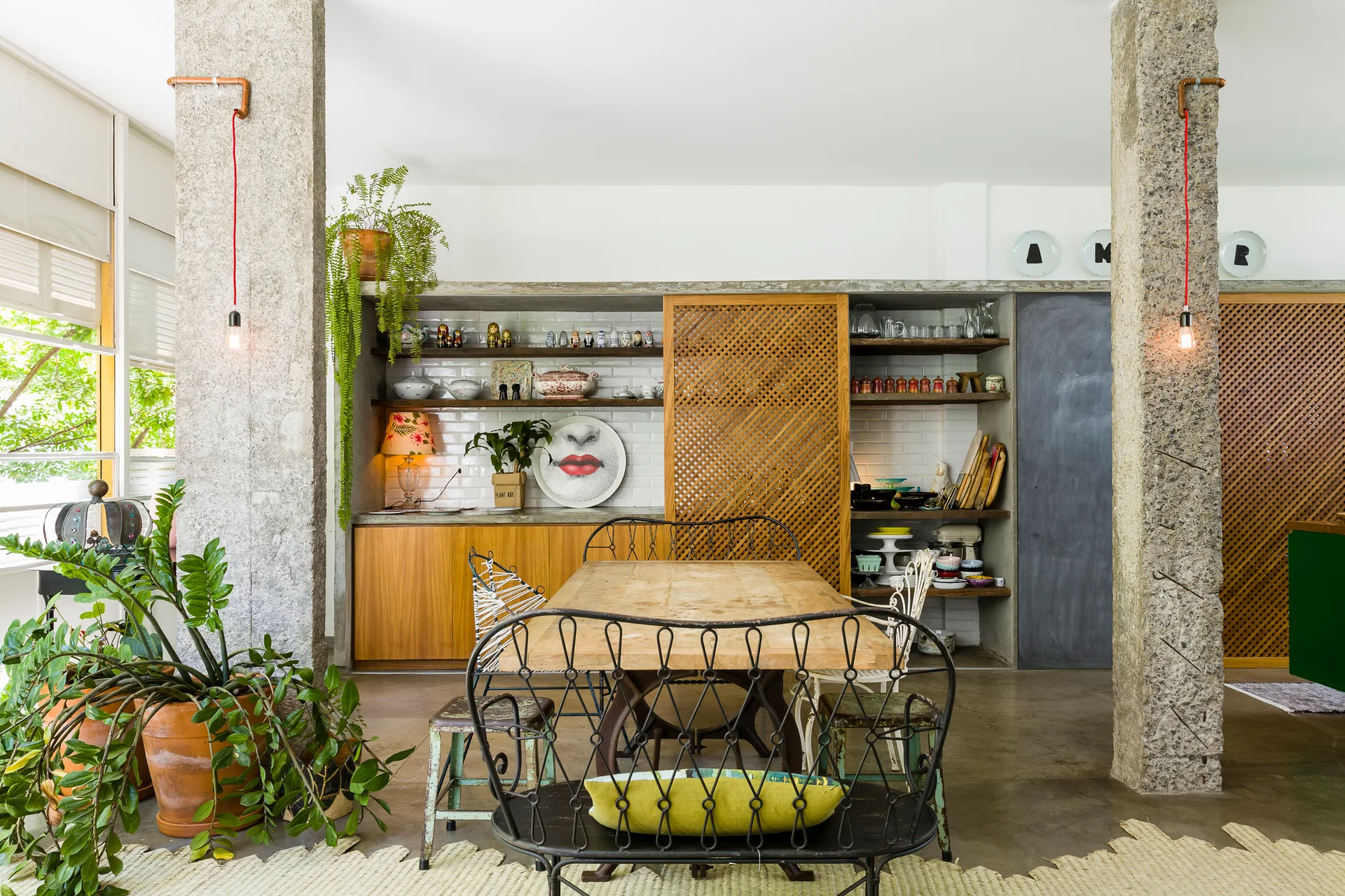 A dining room features a rustic wooden table surrounded by eclectic chairs, set between two rough concrete pillars with a background of lattice sliding doors.