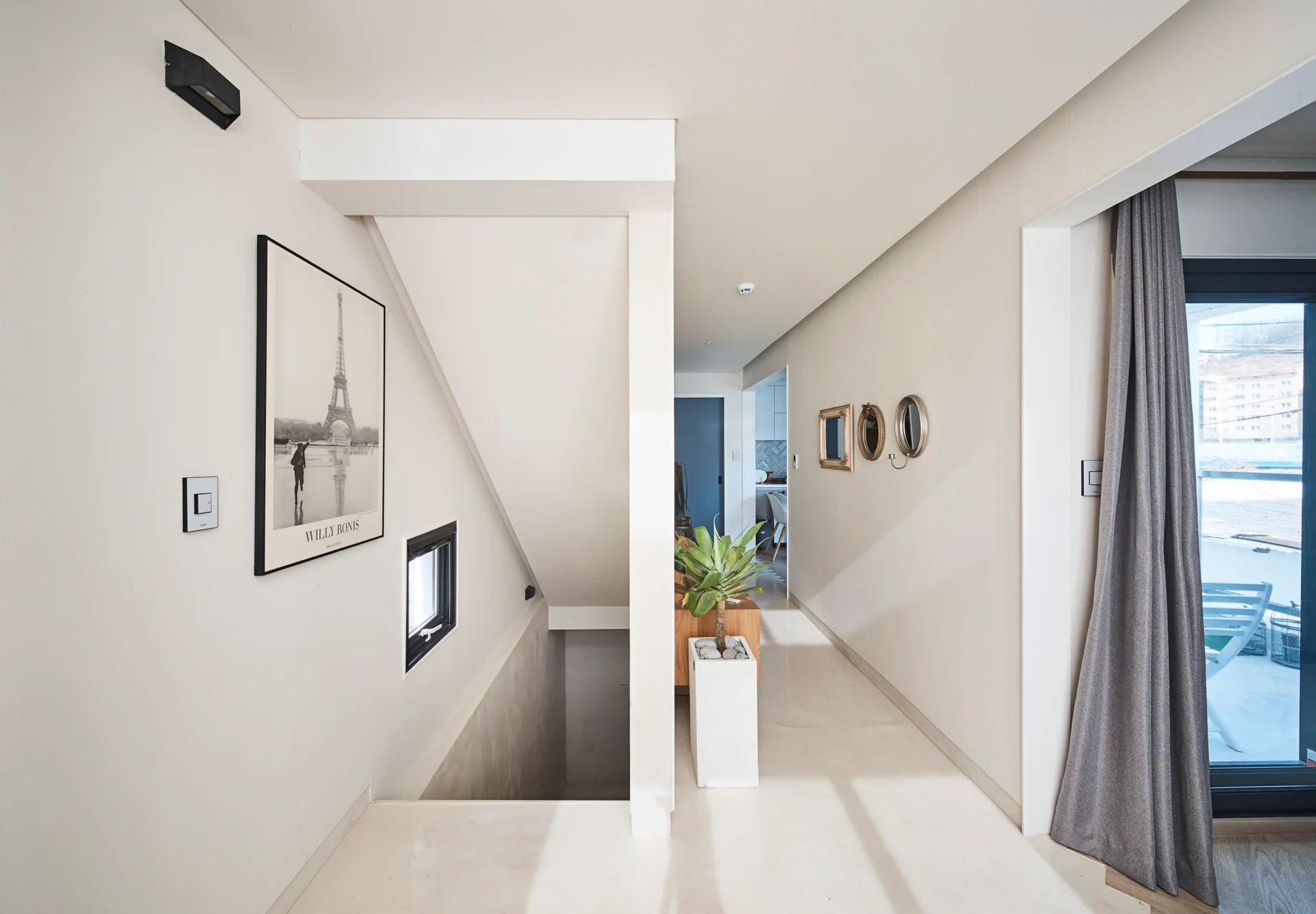 A bright off-white hallway featuring a staircase, a tall white planter with a green plant, and small wall mirrors beside a curtained door.